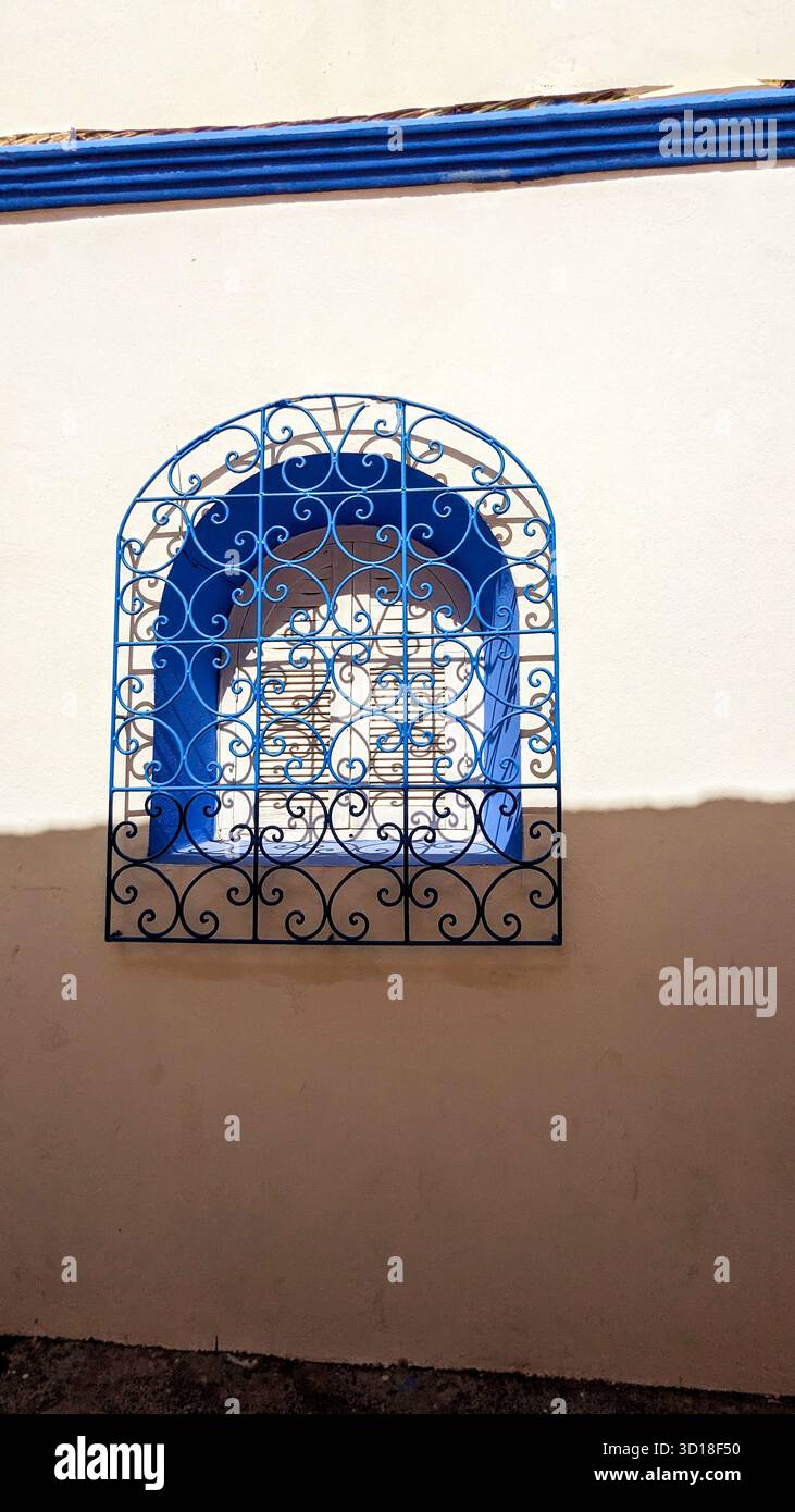 finestra di nicchia blu vibrante con griglia in ferro battuto ornata contro una parete bianca illuminata dal sole e un'ombra profonda - Immagine stock catturata con smartphone