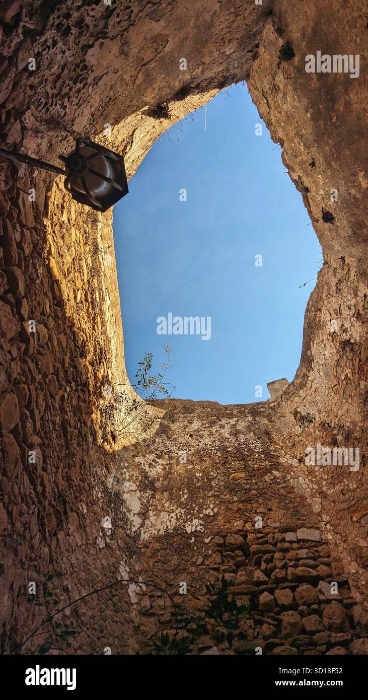 Vista verso l'alto attraverso le rovine circolari di un'antica torre in pietra, che rivela un cielo blu brillante. la luce calda del sole migliora la consistenza delle pareti invecchiate Foto Stock