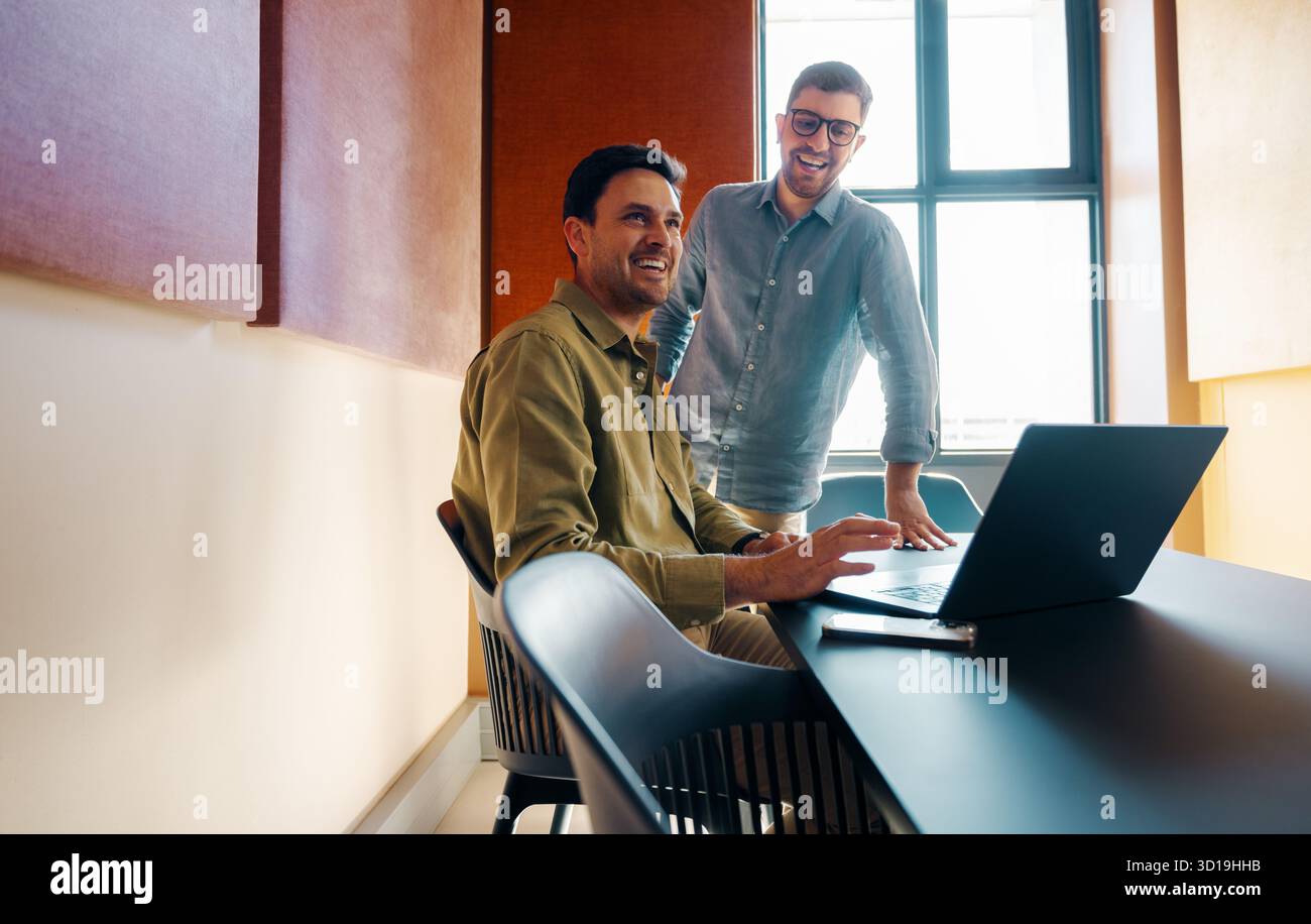 Due uomini di medie dimensioni collaborano comodamente a una scrivania, utilizzando un computer portatile in un ufficio moderno e luminoso che riflette il lavoro di squadra e la creatività Foto Stock