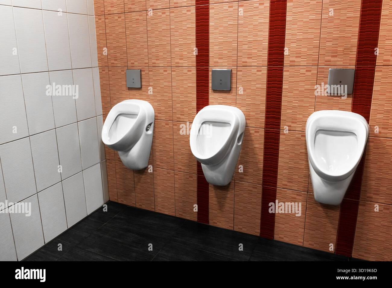 Tre orinatoi bianchi in un bagno pubblico con pareti piastrellate e pavimento scuro. Foto di alta qualità Foto Stock