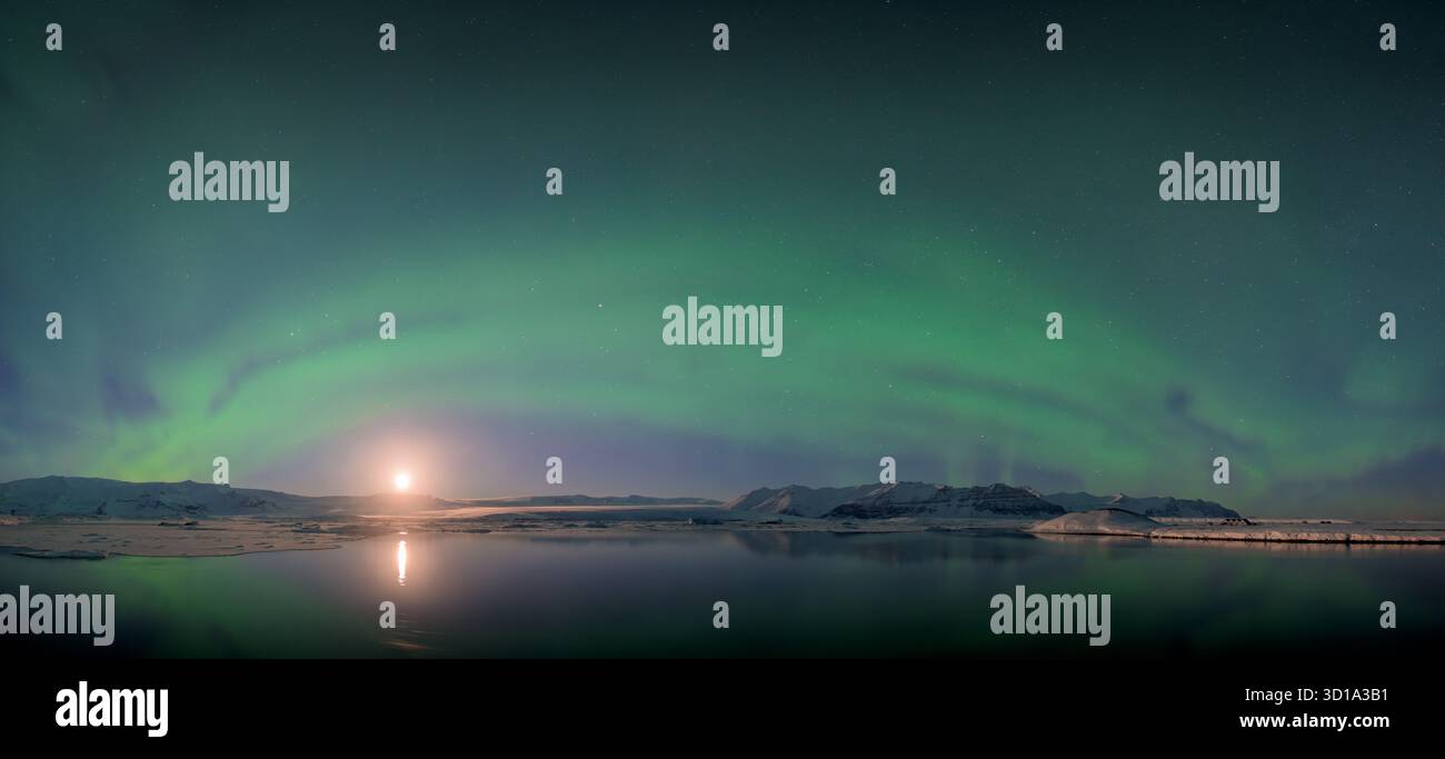 La vista dell'eterea aurora boreale verde danza sopra le acque tranquille che riflettono il soffice bagliore della luna, Jokulsarlon, Sveitarfélagið Hornafjörður, Islanda. Foto Stock