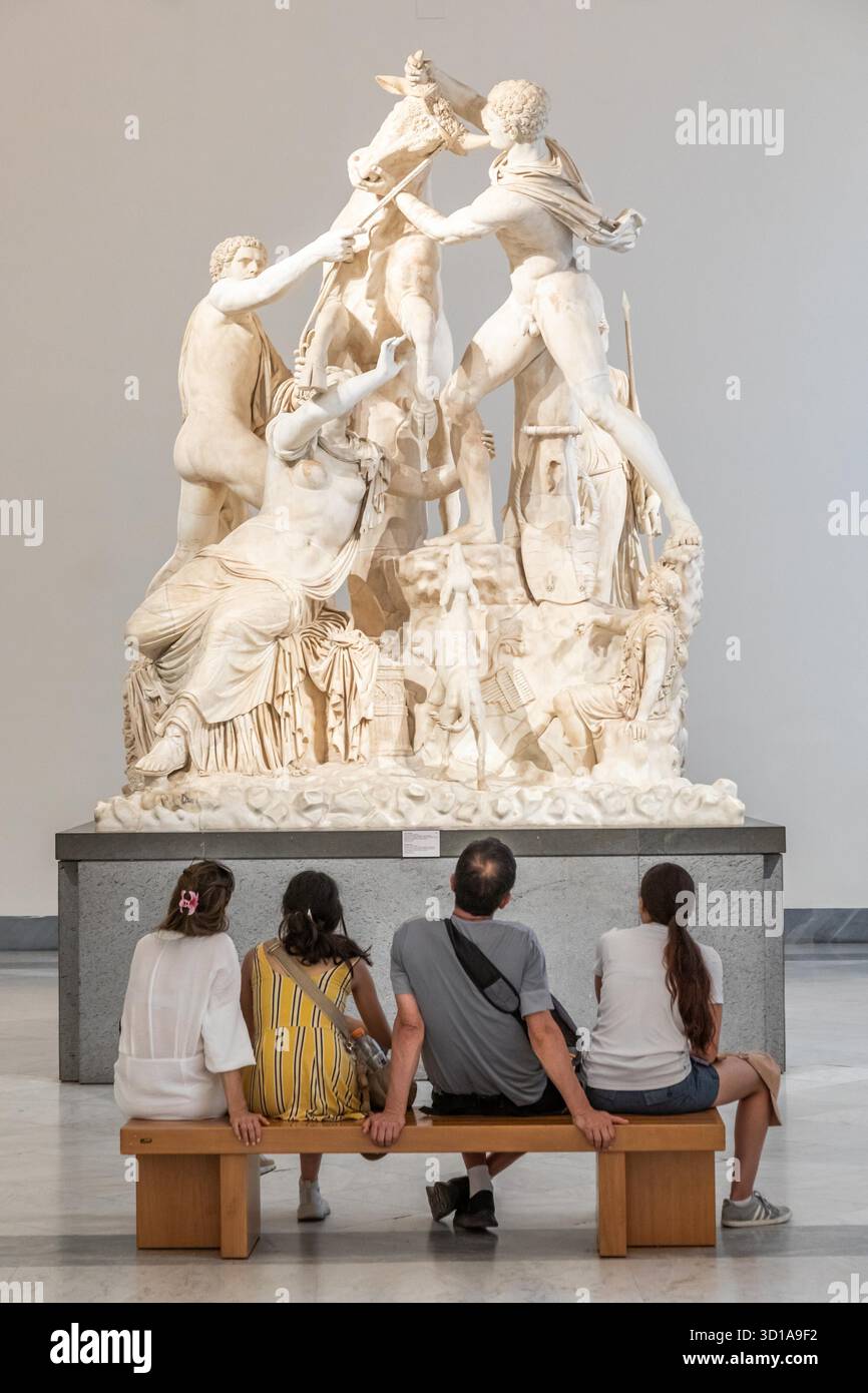 NAPOLI, ITALIA? AGOSTO 2025: Una famiglia siede su una panchina che guarda la colossale statua in marmo del toro Farnese all'interno del Museo Archeologico di Napoli - turismo culturale e arte classica Foto Stock