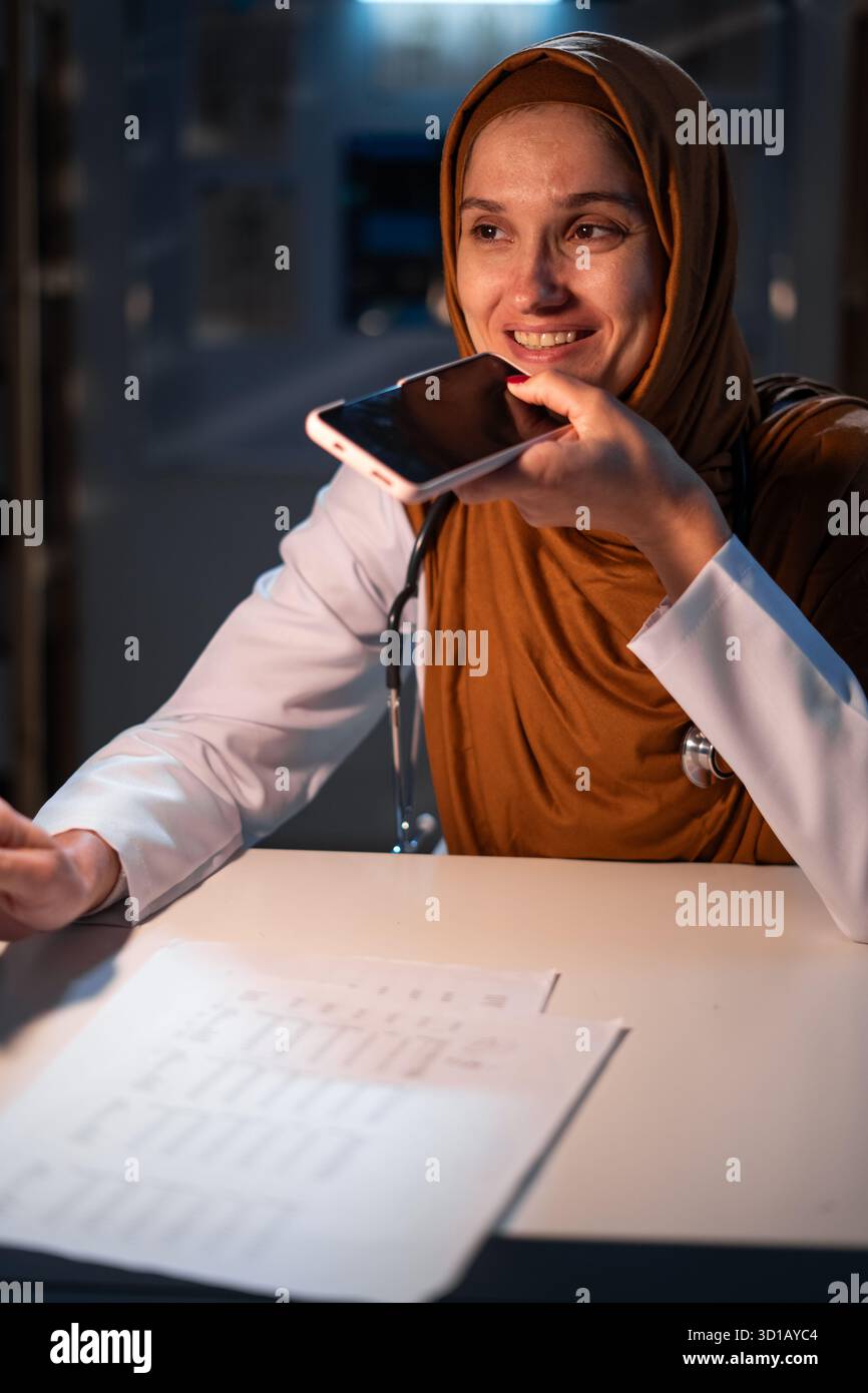 Concetto di intelligenza artificiale in medicina. Medico musulmano in hijab comando ad assistente ai, dettare il messaggio audio utilizzando lo smartphone lavorando su vicino Foto Stock