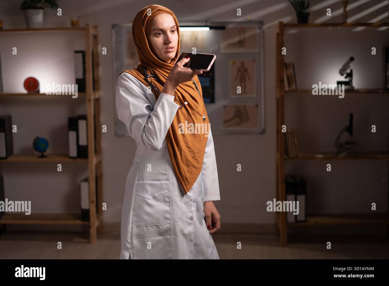 Concetto di intelligenza artificiale in medicina. Medico musulmano in hijab comando ad assistente ai, dettare il messaggio audio utilizzando lo smartphone lavorando su vicino Foto Stock