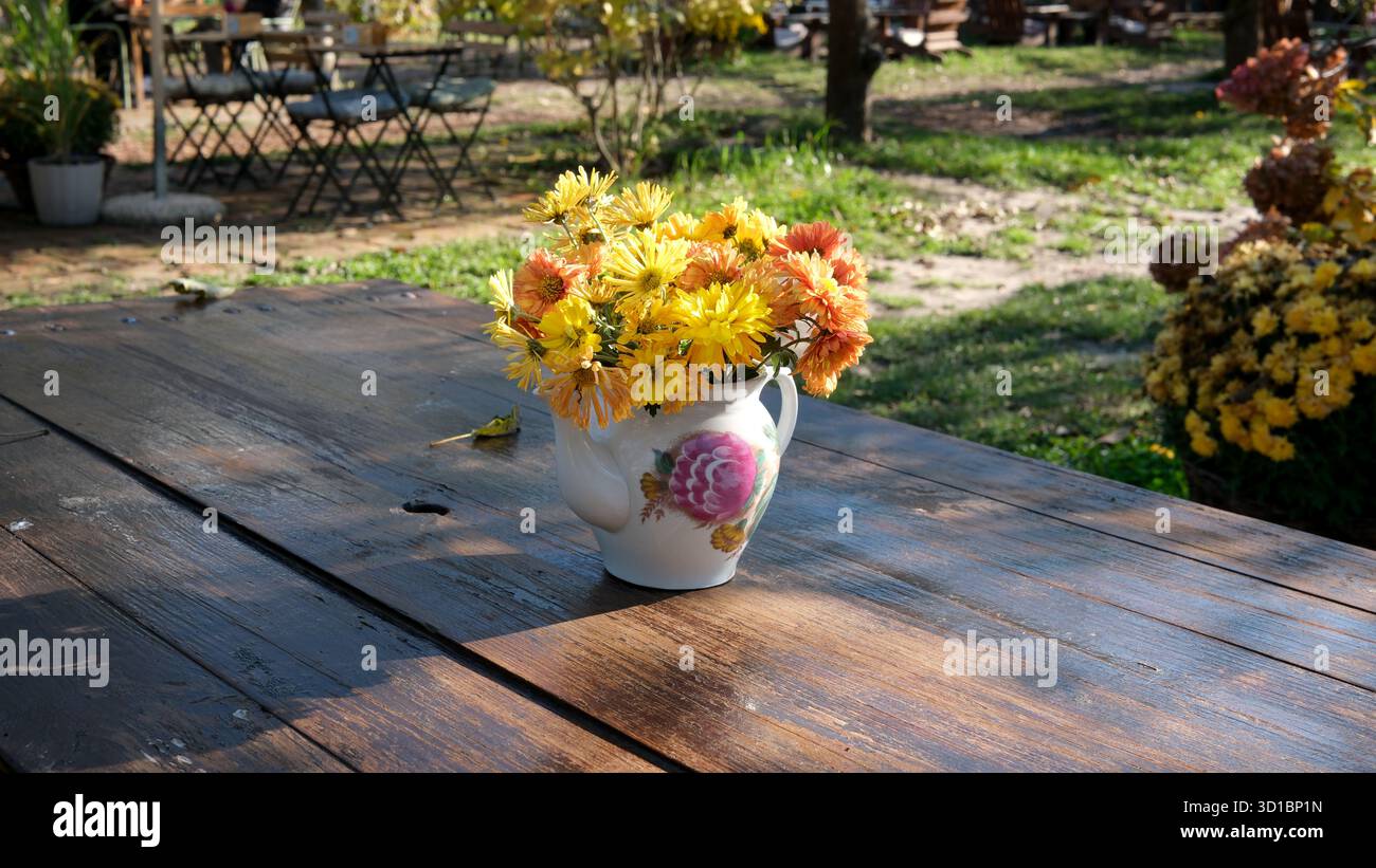 Fiori colorati riposano in un vaso su un tavolo di legno circondato dal verde. Foto Stock