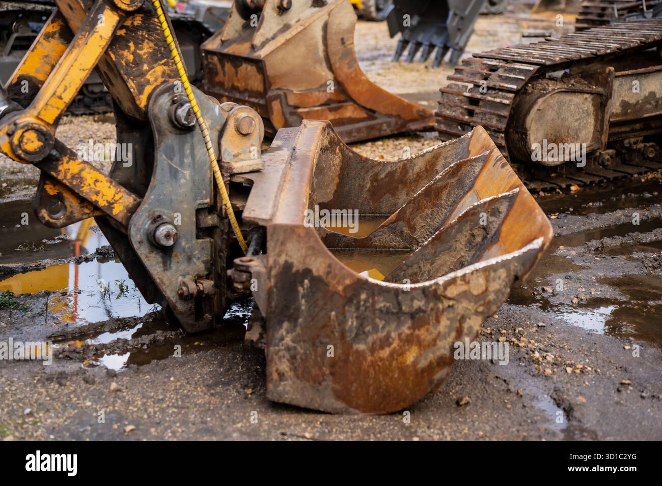 Primo piano della vecchia benna dell'escavatore fissata al braccio idraulico in un cantiere fangoso che mostra ruggine, usura e dettagli meccanici dei macchinari pesanti utilizzati Foto Stock