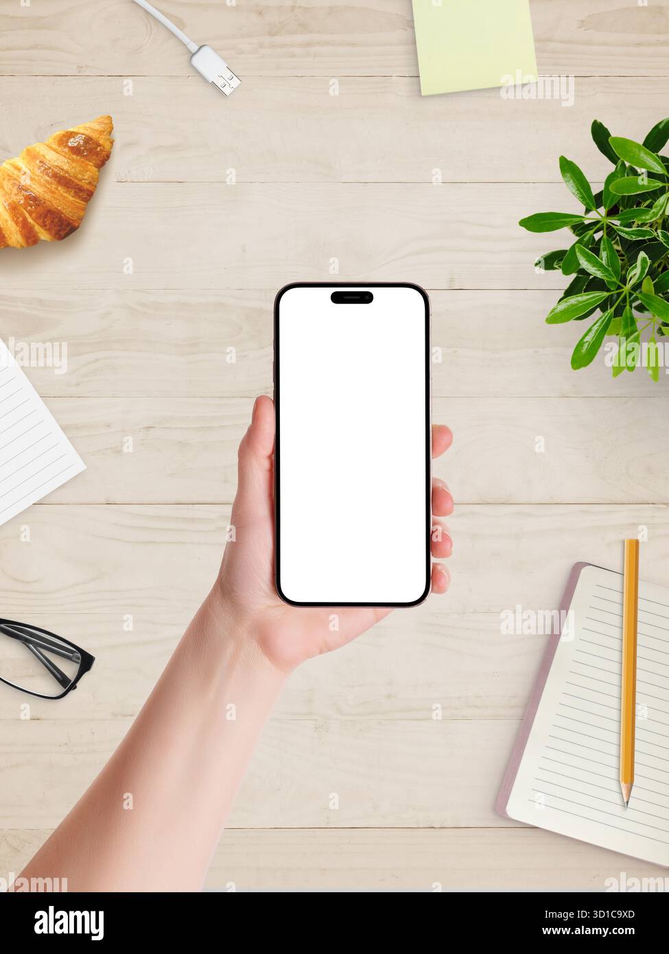 Mano che tiene lo smartphone con schermo bianco vuoto sopra la scrivania, croissant, notebook e impianto, pronto per il branding o il design mobile Foto Stock