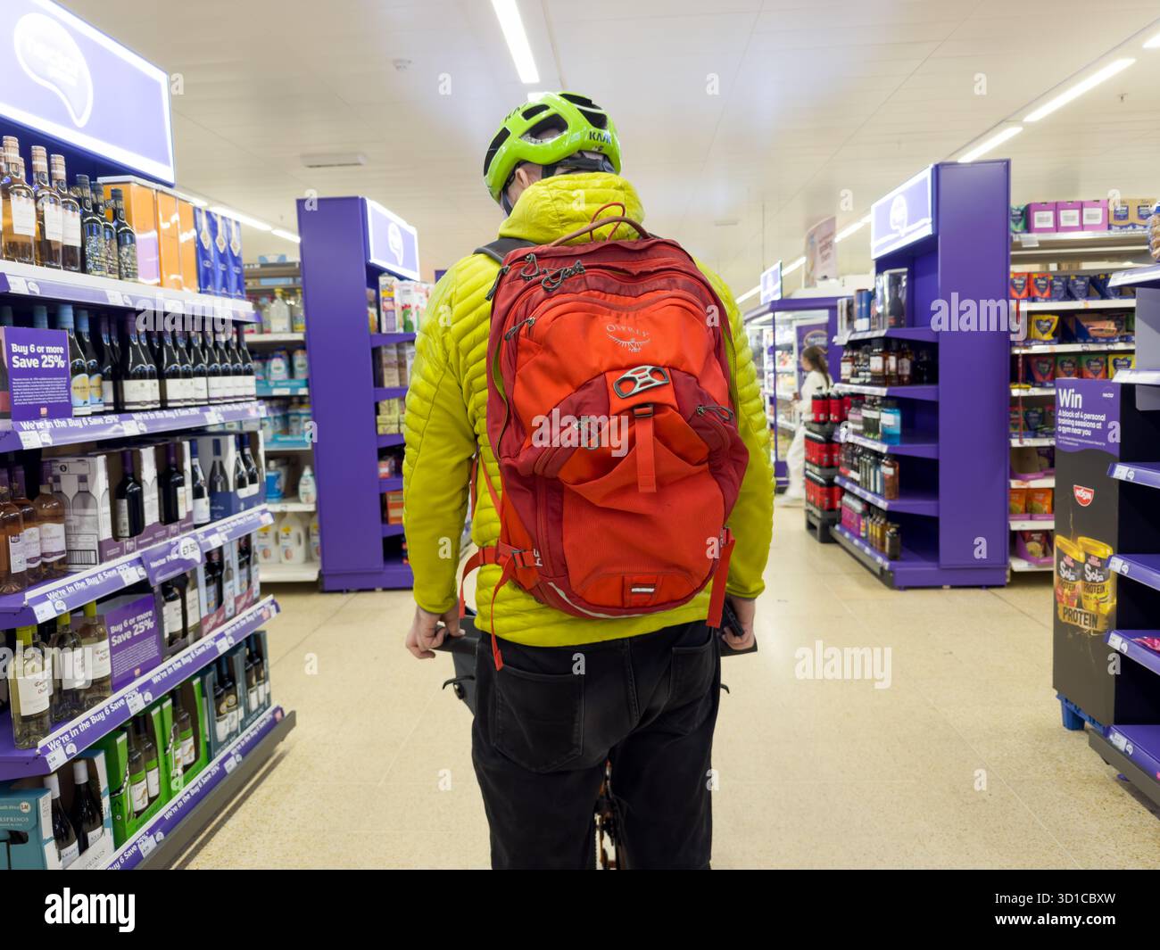 Un ciclista vestito da una giacca gialla con pelo, uno zaino rosso e un casco verde da bici, esplora la sala del vino all'interno di un supermercato di Londra nel regno unito Foto Stock