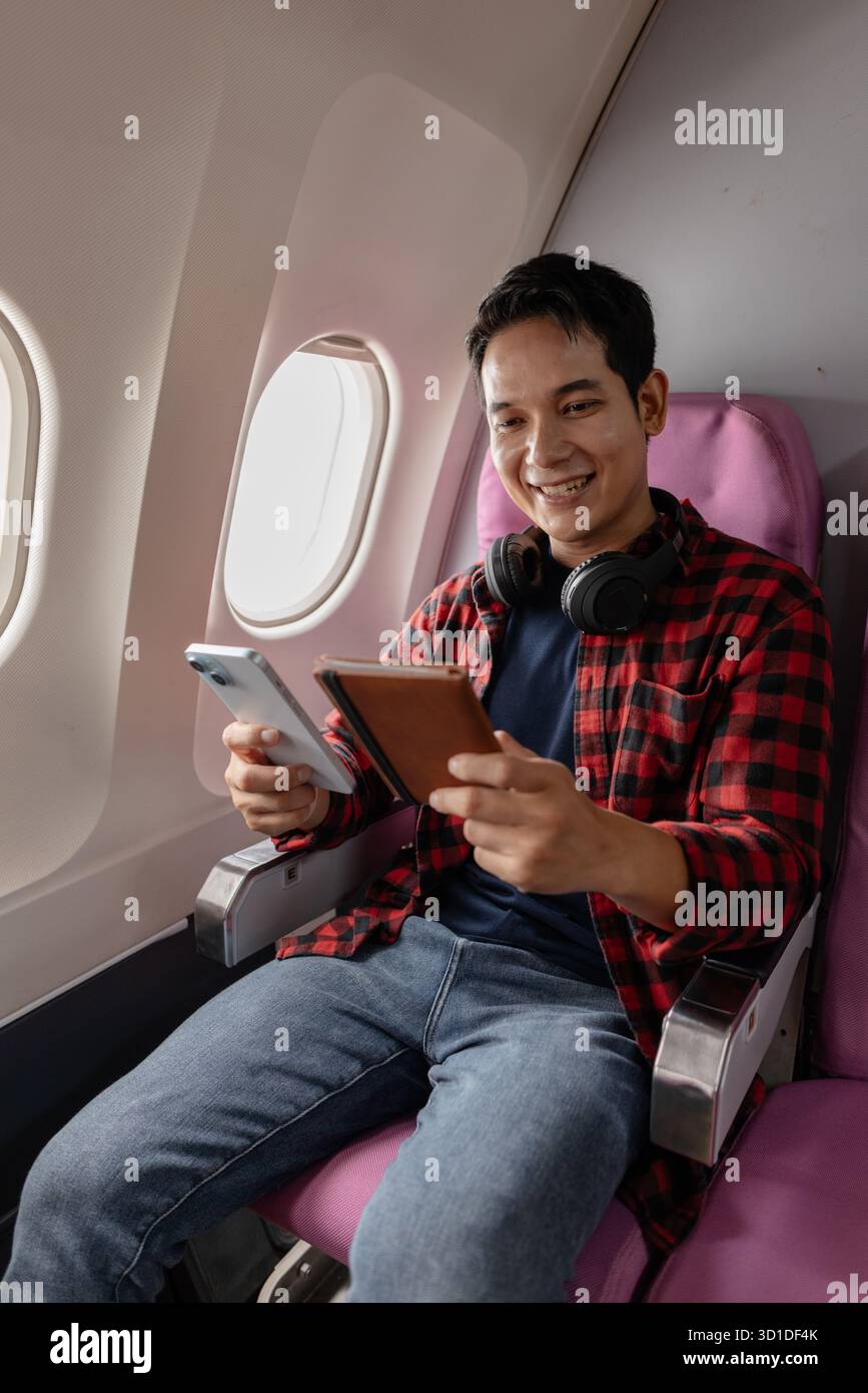 Un giovane uomo siede su un aereo, godendo il volo mentre controlla il suo smartphone e il passaporto. Sembra rilassato e contento, riflettendo il moderno Foto Stock