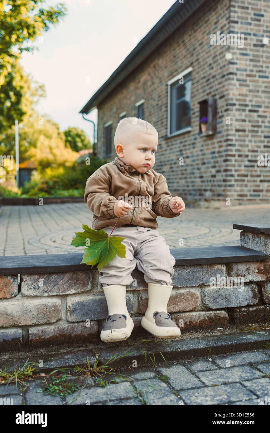 Un bambino gioioso siede su gradini di pietra, tenendo in mano una foglia verde vibrante, simboleggia lo spirito spensierato e avventuroso dell'infanzia in un natur colorato e vivace Foto Stock