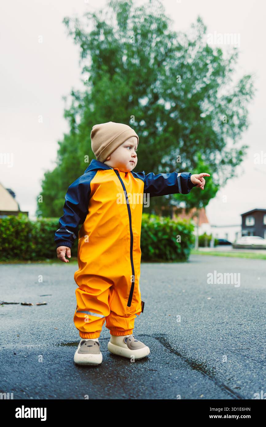 In una giornata di pioggia, un bambino gioioso con tuta da pioggia gialla e cappello in maglia indica qualcosa sul marciapiede, incarnando la curiosità dell'infanzia e la gioia di un'esperienza expl Foto Stock