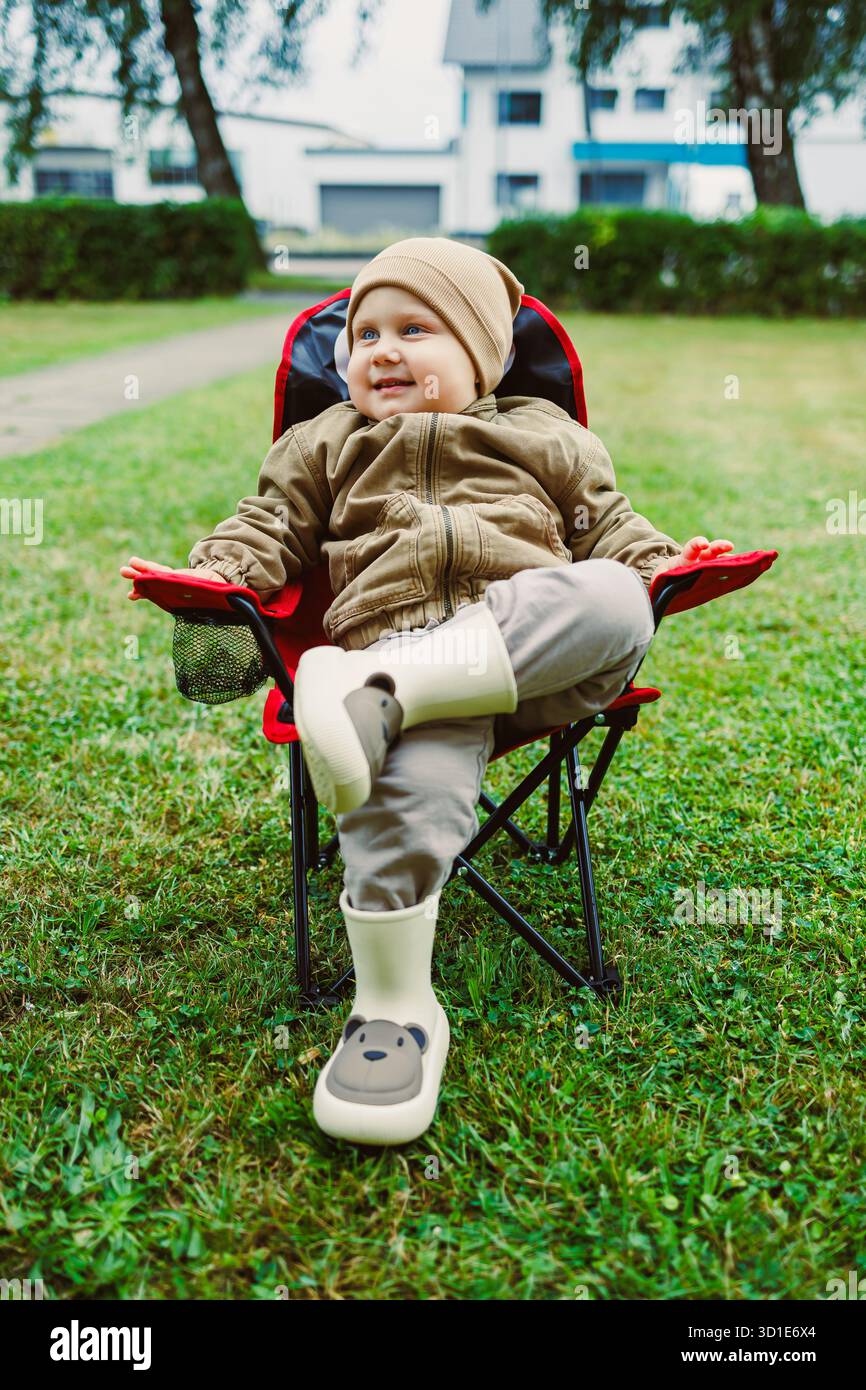 Un bambino allegro in un abito elegante siede su una piccola sedia in un vivace prato verde, catturando un momento delizioso perfetto per il divertimento all'aria aperta in famiglia e un Foto Stock