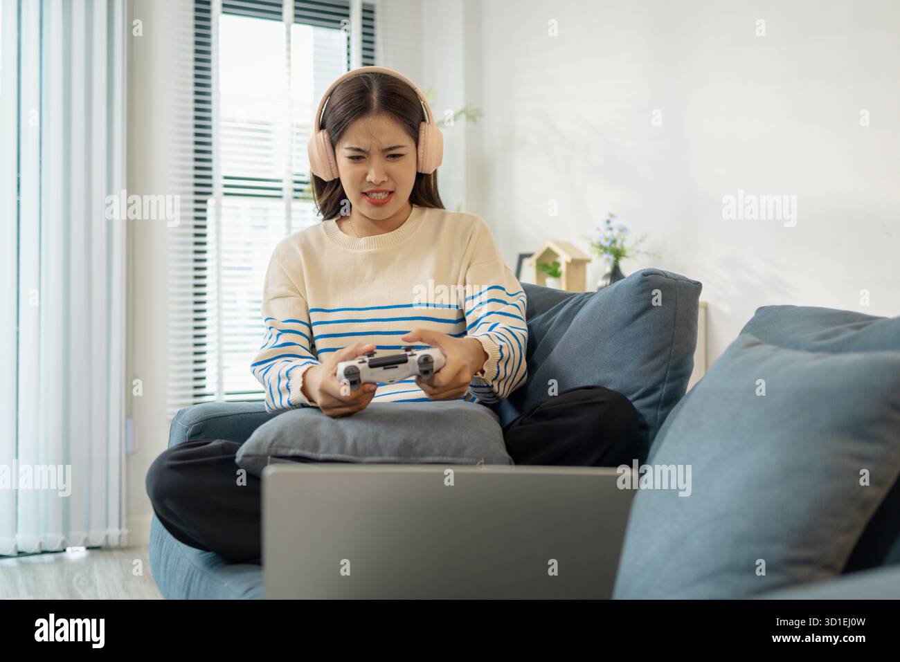 Una giovane donna che indossa le cuffie sta giocando a un videogame sul suo notebook, tenendo in mano un controller con espressioni emotive di gioia, eccitazione e fruste Foto Stock