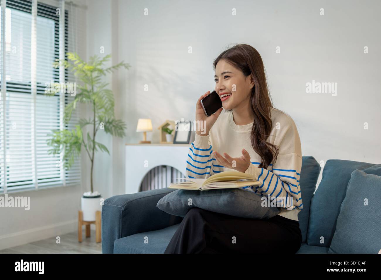 Una giovane donna seduta comodamente su un divano, sorridente mentre utilizza uno smartphone con un notebook in grembo, mostrando una scena di multitasking tra digi Foto Stock