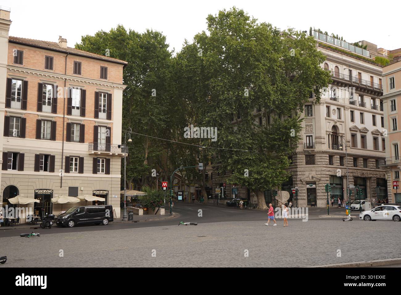 Ampia piazza lastricata dominata da grandi alberi, caratterizzata da edifici romani classici e da persone che camminano lungo la piazza vicino a un ingresso della metropolitana. Foto Stock