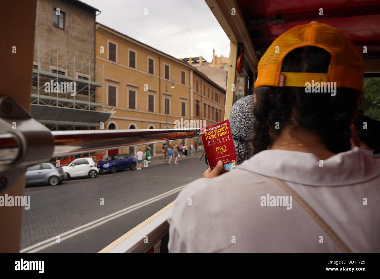 Vista sulla strada dalla vetrina di un autobus turistico di Roma, con edifici storici in ocra, auto parcheggiate e un turista con una guida turistica "Big Bus Tours". Foto Stock