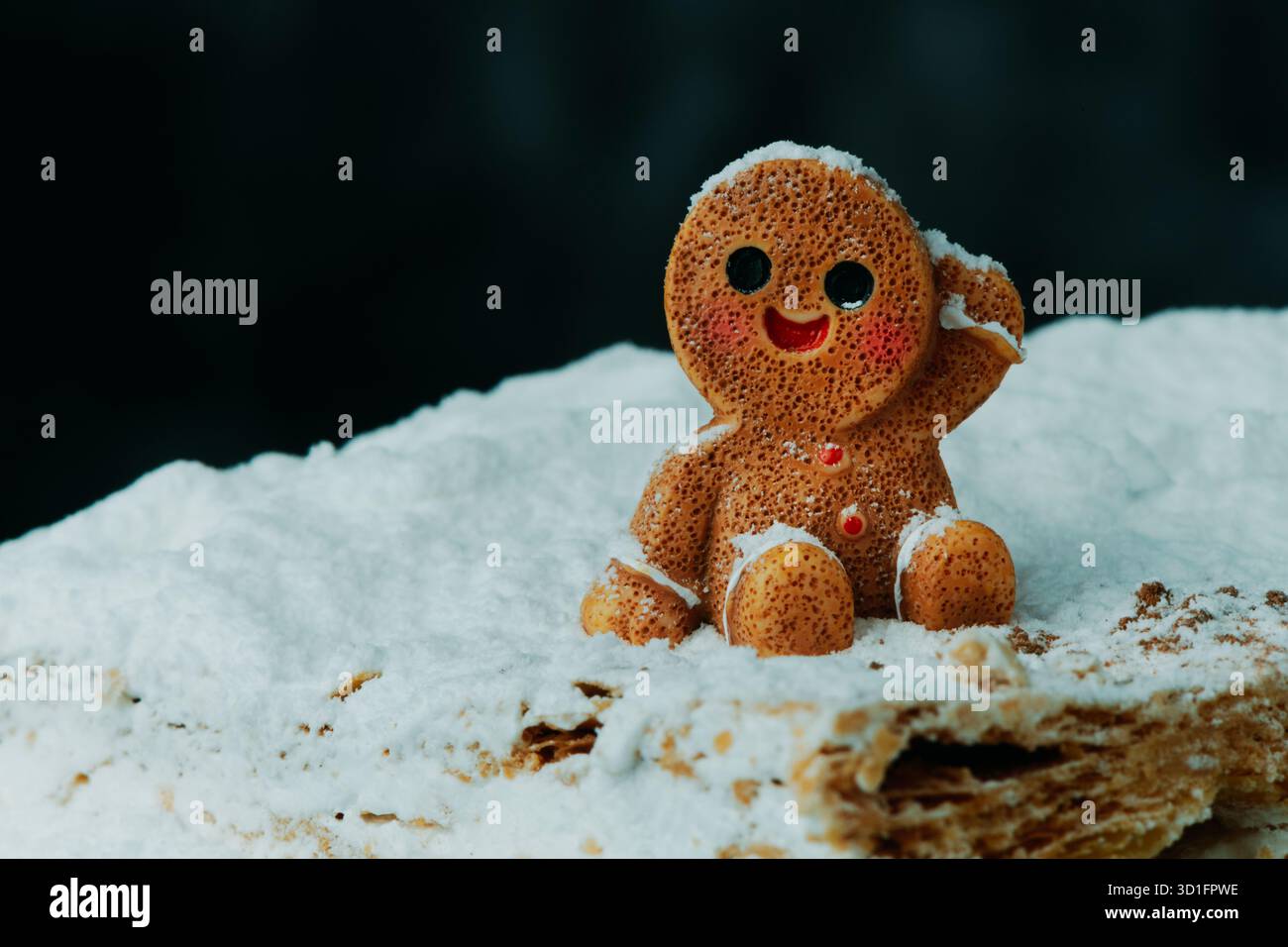 un piccolo uomo di pan di zenzero si siede sul bordo di una superficie nevosa milhoja, i suoi bottoni rossi brillano attraverso la delicata polvere bianca dello zucchero che cade Foto Stock