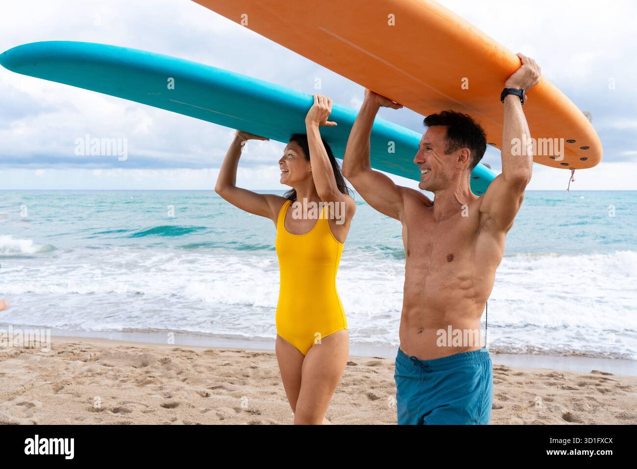 Coppia che cammina su una spiaggia sabbiosa, trasporta tavole da surf sulle spalle, pronta per uno stile di vita attivo e abbraccia la sfida di imparare una nuova acqua Foto Stock