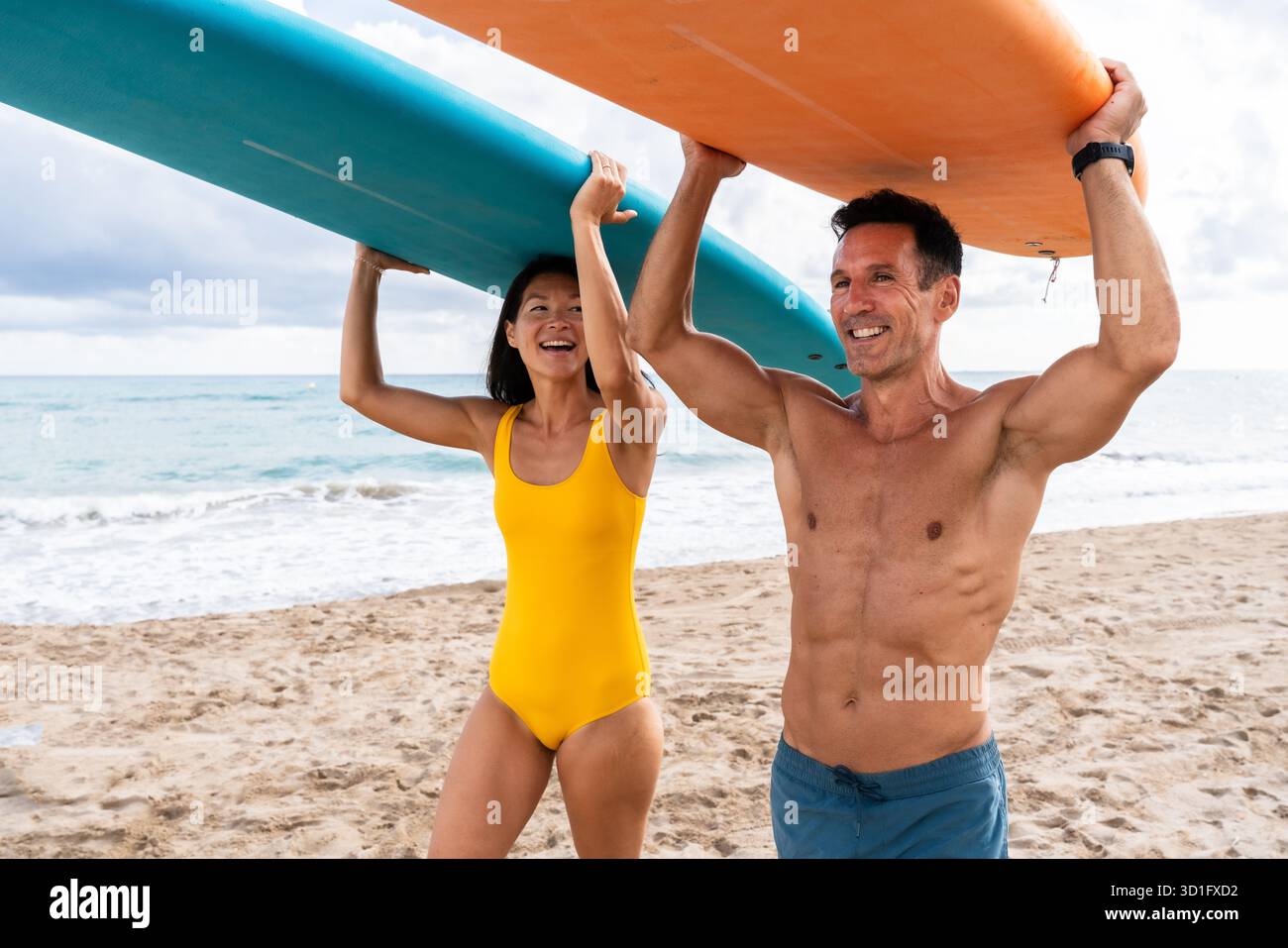 Coppia che cammina su una spiaggia sabbiosa, trasporta tavole da surf sulle spalle, pronta per uno stile di vita attivo e abbraccia la sfida di imparare una nuova acqua Foto Stock