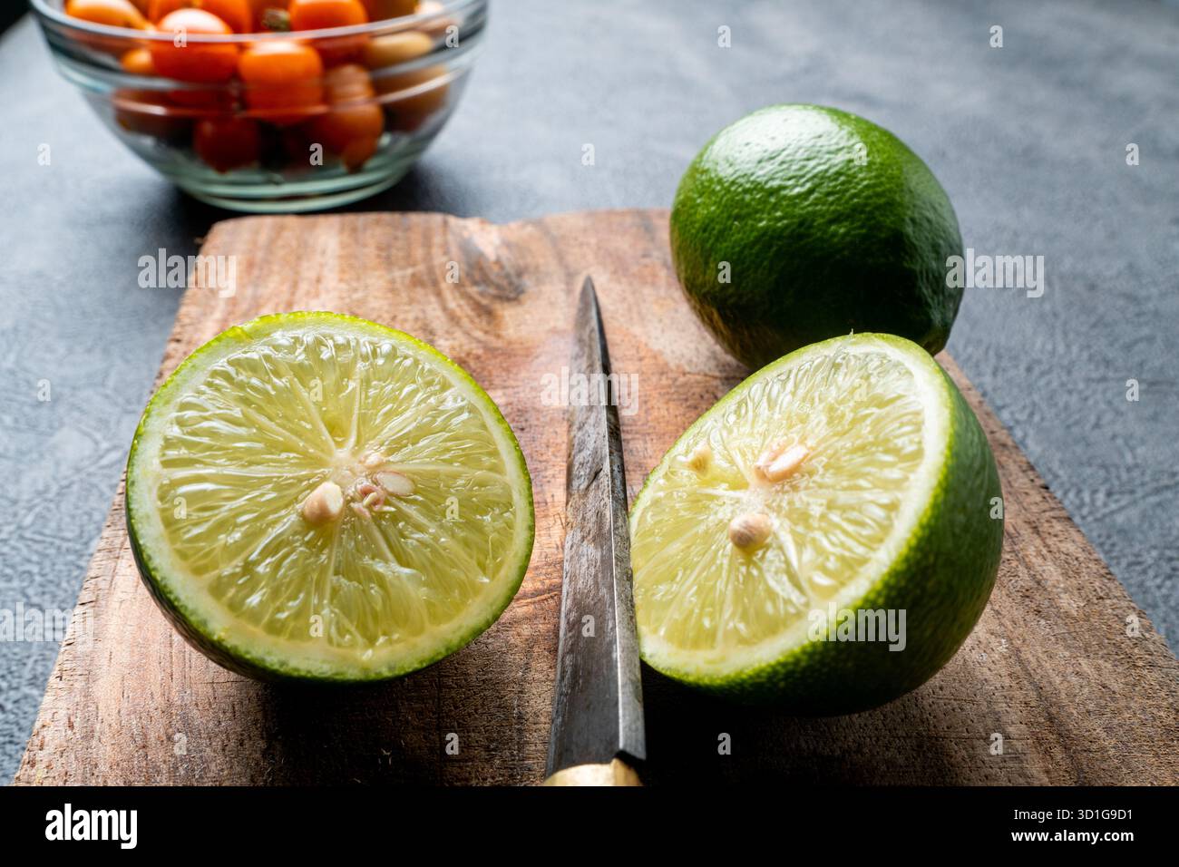 Primo piano di limoni verdi freschi, interi e tagliati a fette. Gli agrumi si trovano su un tagliere in legno rustico con uno sfondo scuro e ruvido. Foto Stock