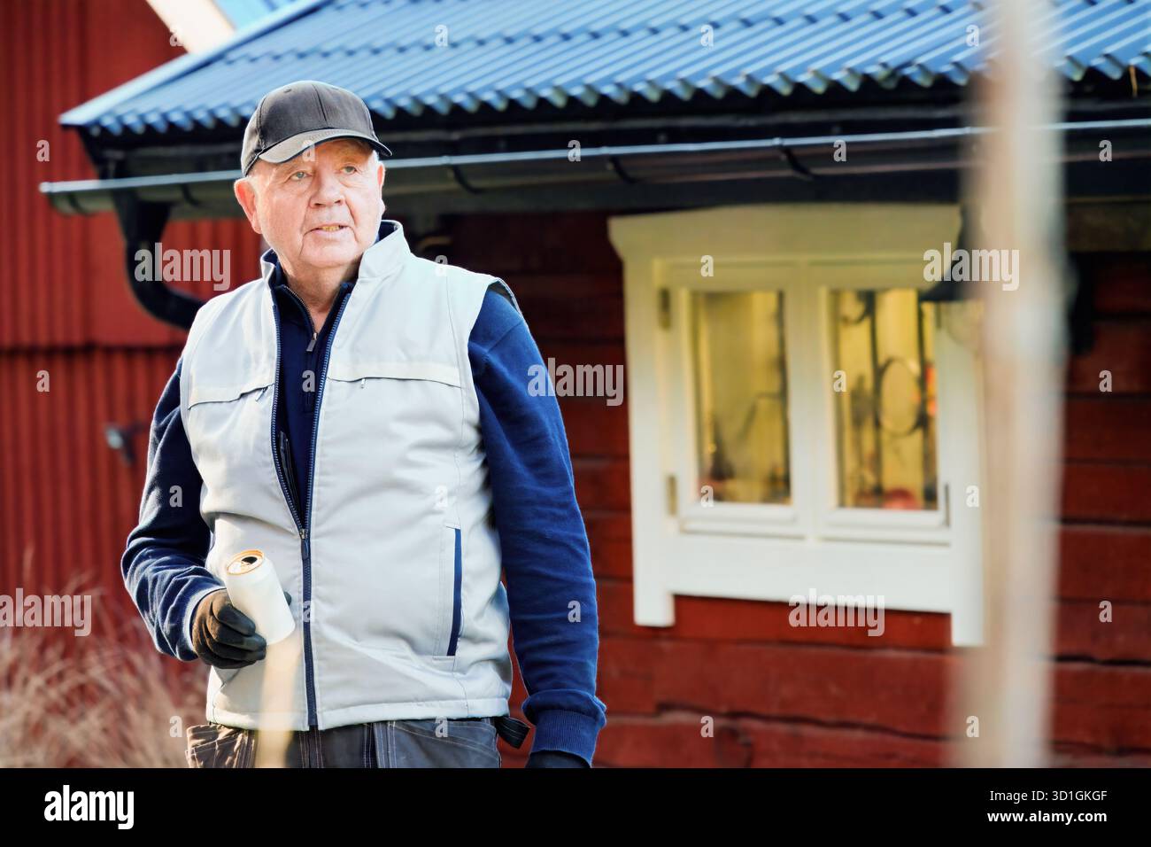 L'uomo anziano in un giubbotto e cappellino è fuori da una casa di legno, in possesso di un barattolo di vernice, pronto per lavori di miglioramento domestico in un ambiente tranquillo all'aperto Foto Stock