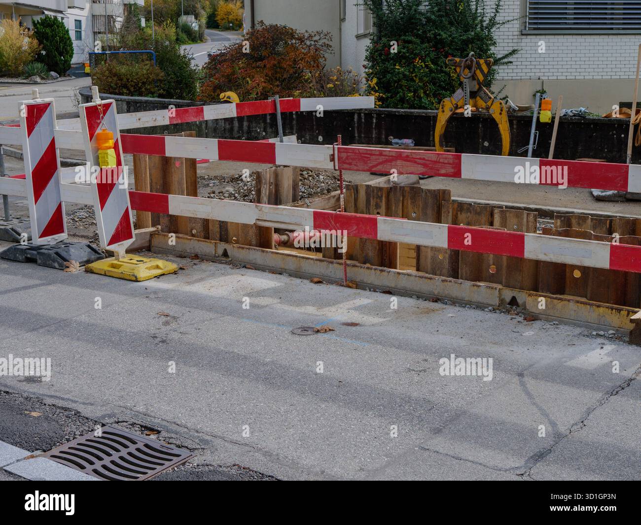 Cantiere stradale urbano con barriere di sicurezza rosse e bianche e escavatore che lavora nell'area di manutenzione stradale. Foto di alta qualità Foto Stock