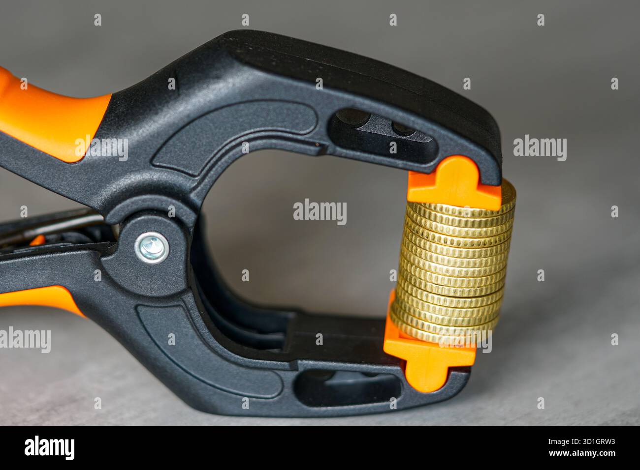 Primo piano di monete d'oro strette in un morsetto nero e arancione, simboleggiando la pressione finanziaria, l'inflazione, la crisi economica e lo stress monetario Foto Stock