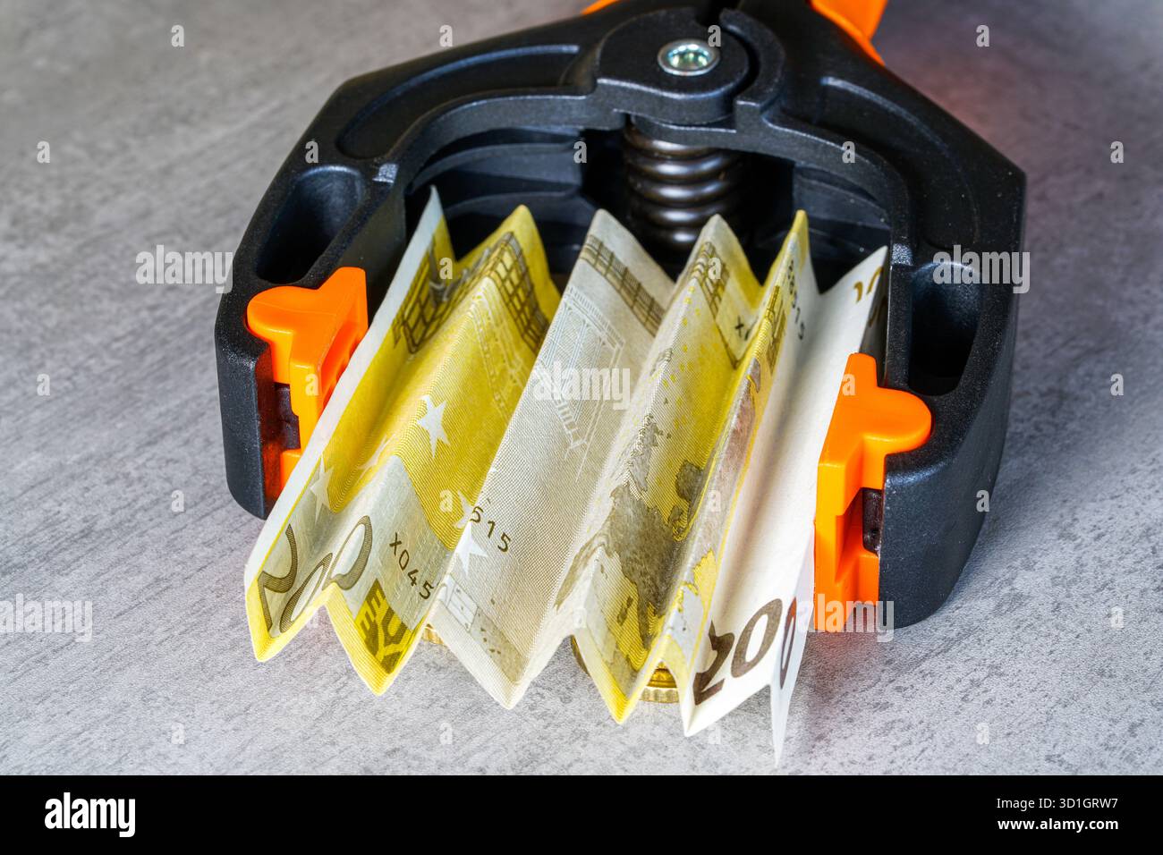 Primo piano delle banconote in euro strettamente schiacciate in un morsetto nero e arancione, simboleggiando la pressione finanziaria, l’inflazione, la crisi economica e il costo della vita Foto Stock