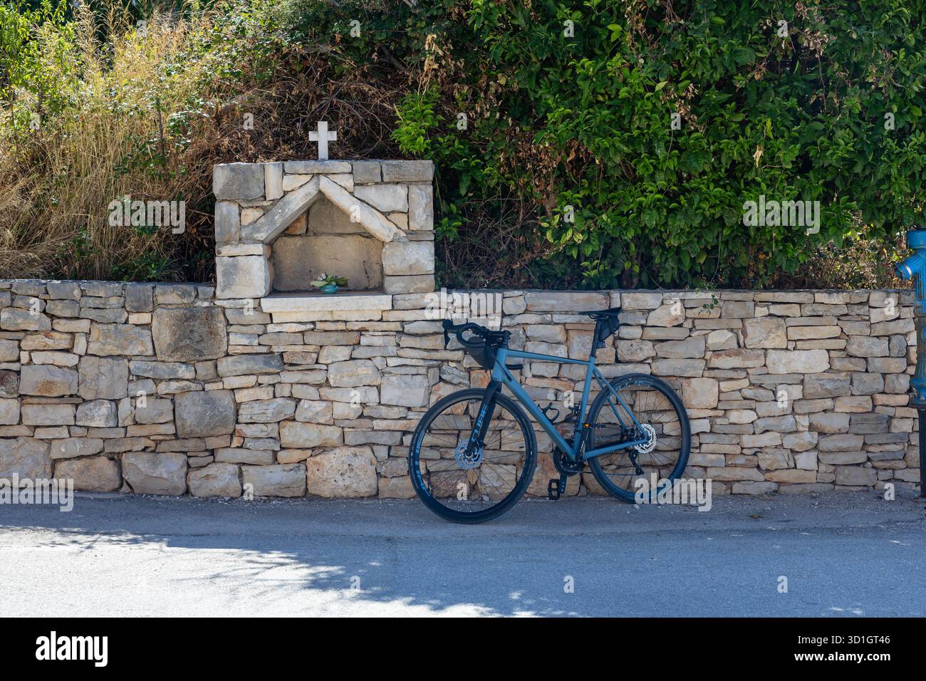SOLTA, CROAZIA, 08.31.2025: Un'elegante bicicletta blu si appoggia su un rustico muro di pietra accanto a un piccolo santuario con una croce, adagiato su una vegetazione lussureggiante. Questo è Foto Stock