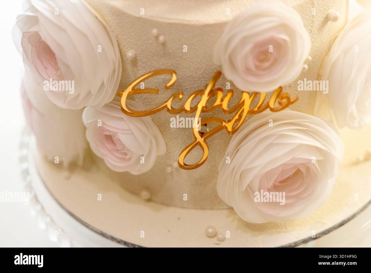 Un'elegante torta adornata da delicate rose bianche e blush, con un'iscrizione dorata ed elegante che recita "??????" ' al centro. Foto Stock