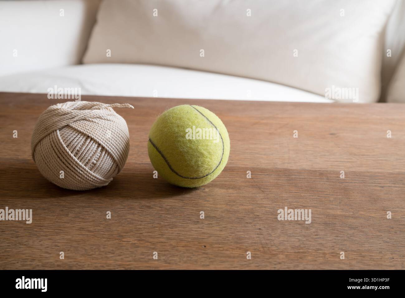 Una palla da tennis verde si trova accanto a una palla di spago beige su un tavolo di legno, con un morbido divano bianco sfocato sullo sfondo. Foto Stock