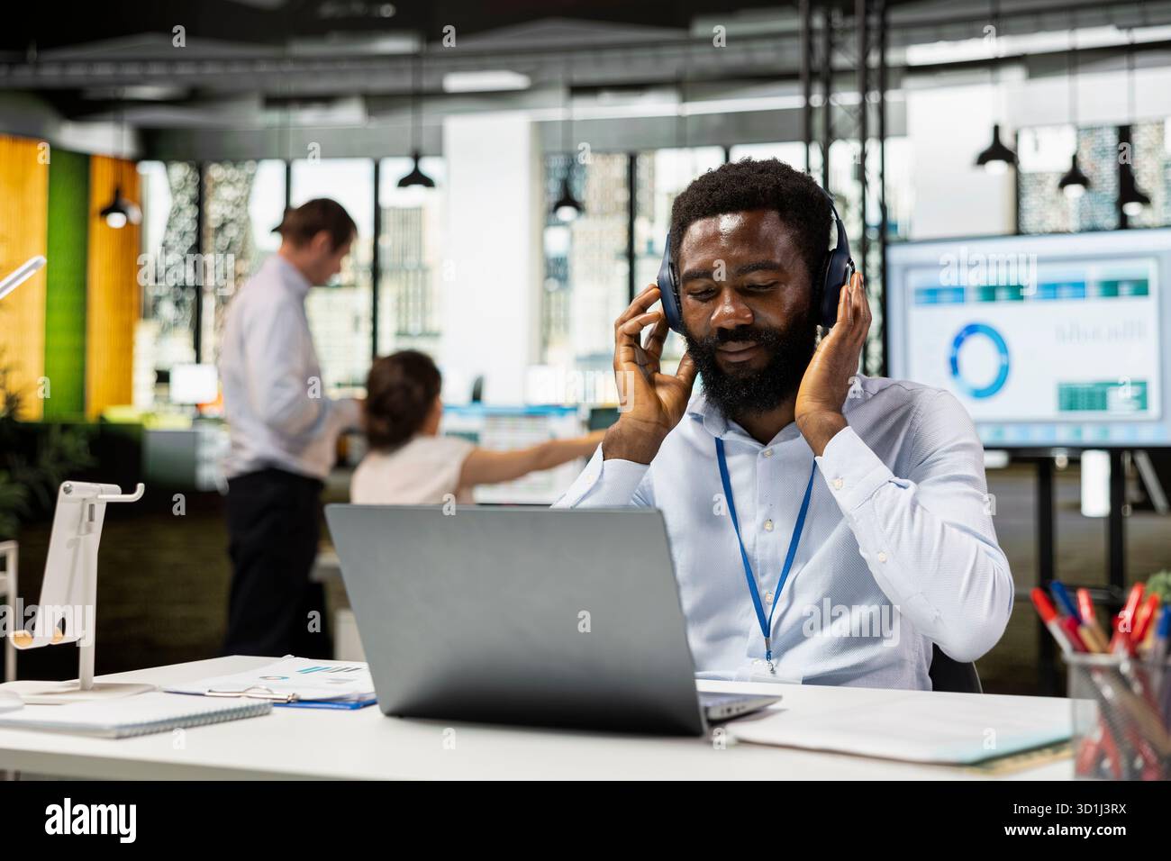 Impiegato che utilizza un computer portatile e ascolta musica, controlla gli aggiornamenti online dell'azienda. L'uomo d'affari tocca le cuffie, ascoltando playlist e finalizzando il report sui fondi di budget nello spazio di lavoro aziendale. Foto Stock