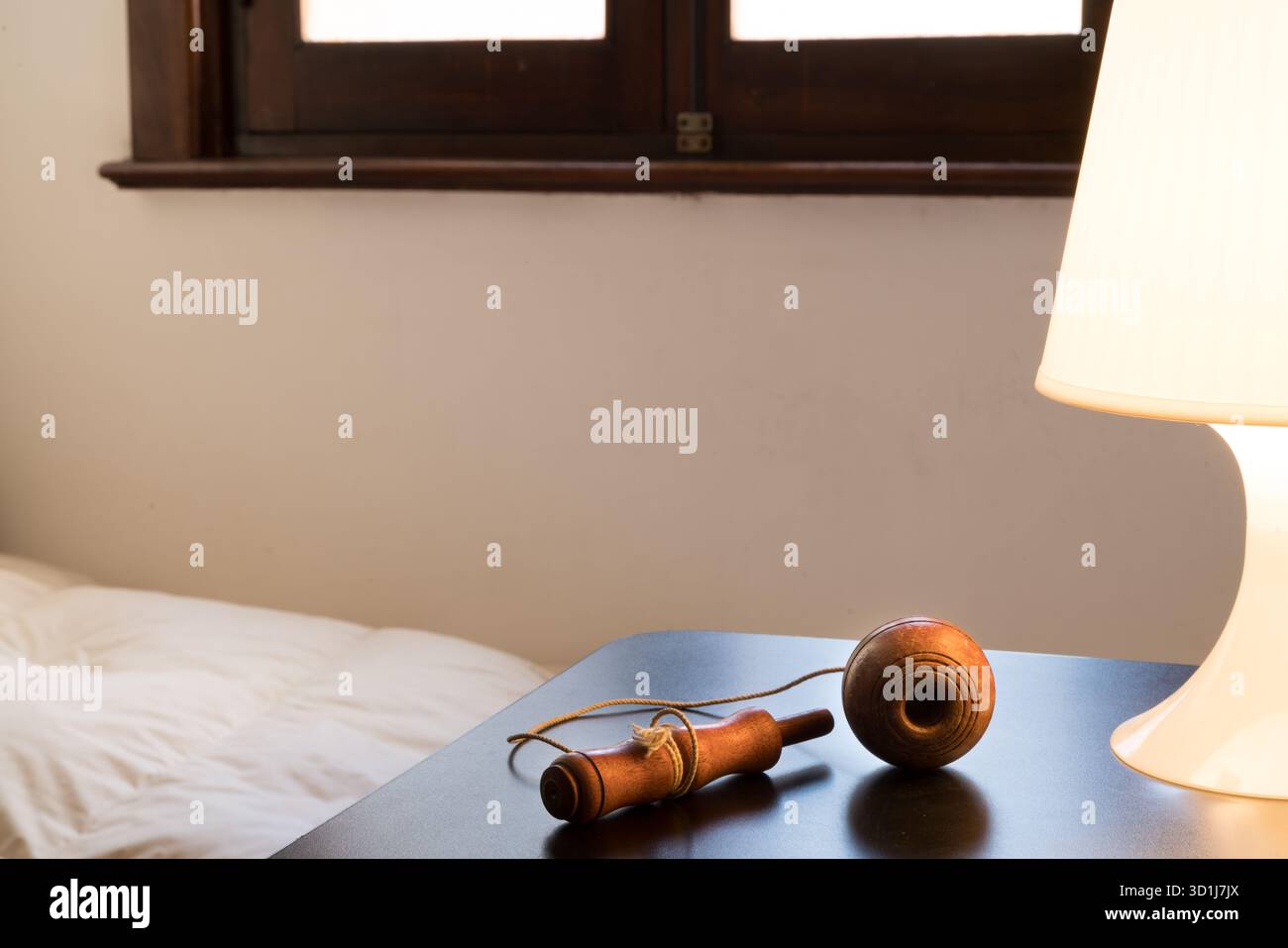 Un tradizionale giocattolo balero in legno poggia su un comodino scuro, illuminato da una lampada bianca brillante, con un letto sfocato e una finestra sullo sfondo. Foto Stock