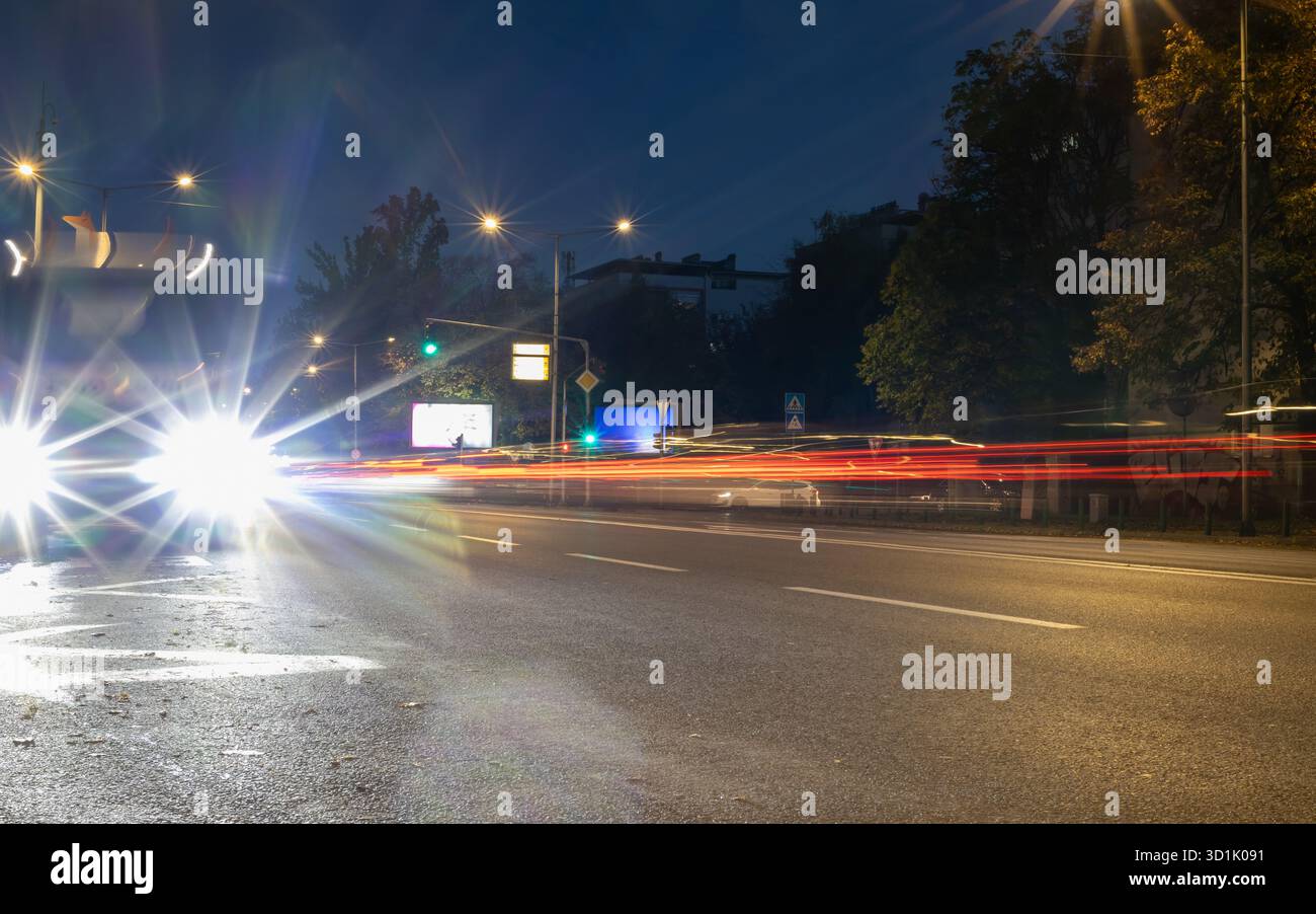 Foto notturne vivide a lunga esposizione di strade cittadine con fari luminosi per auto, tracce luminose di traffico in movimento, lampioni stradali e fogliame scuro Foto Stock