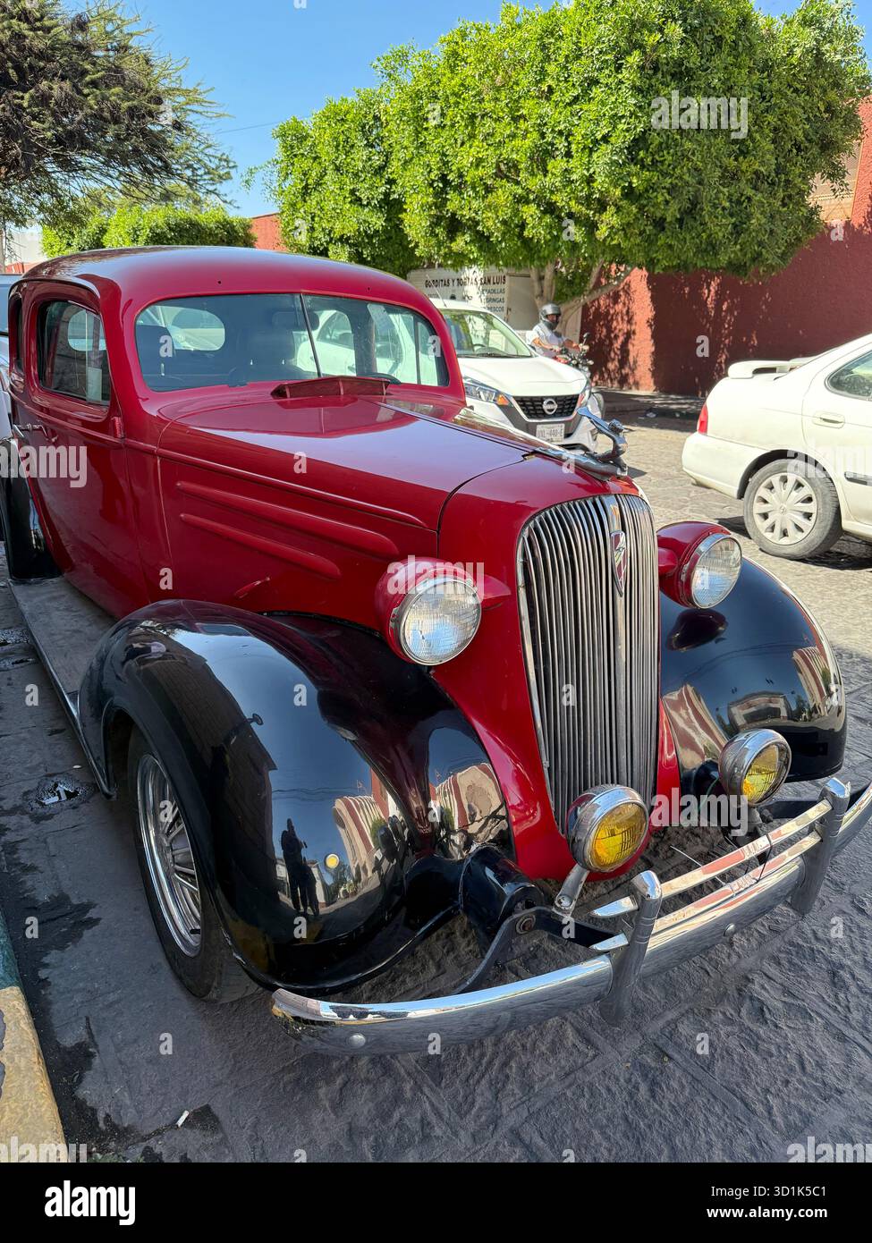 Auto d'epoca rossa e nera parcheggiata su una strada illuminata dal sole con alberi e altri veicoli sullo sfondo - Immagine stock catturata con smartphone
