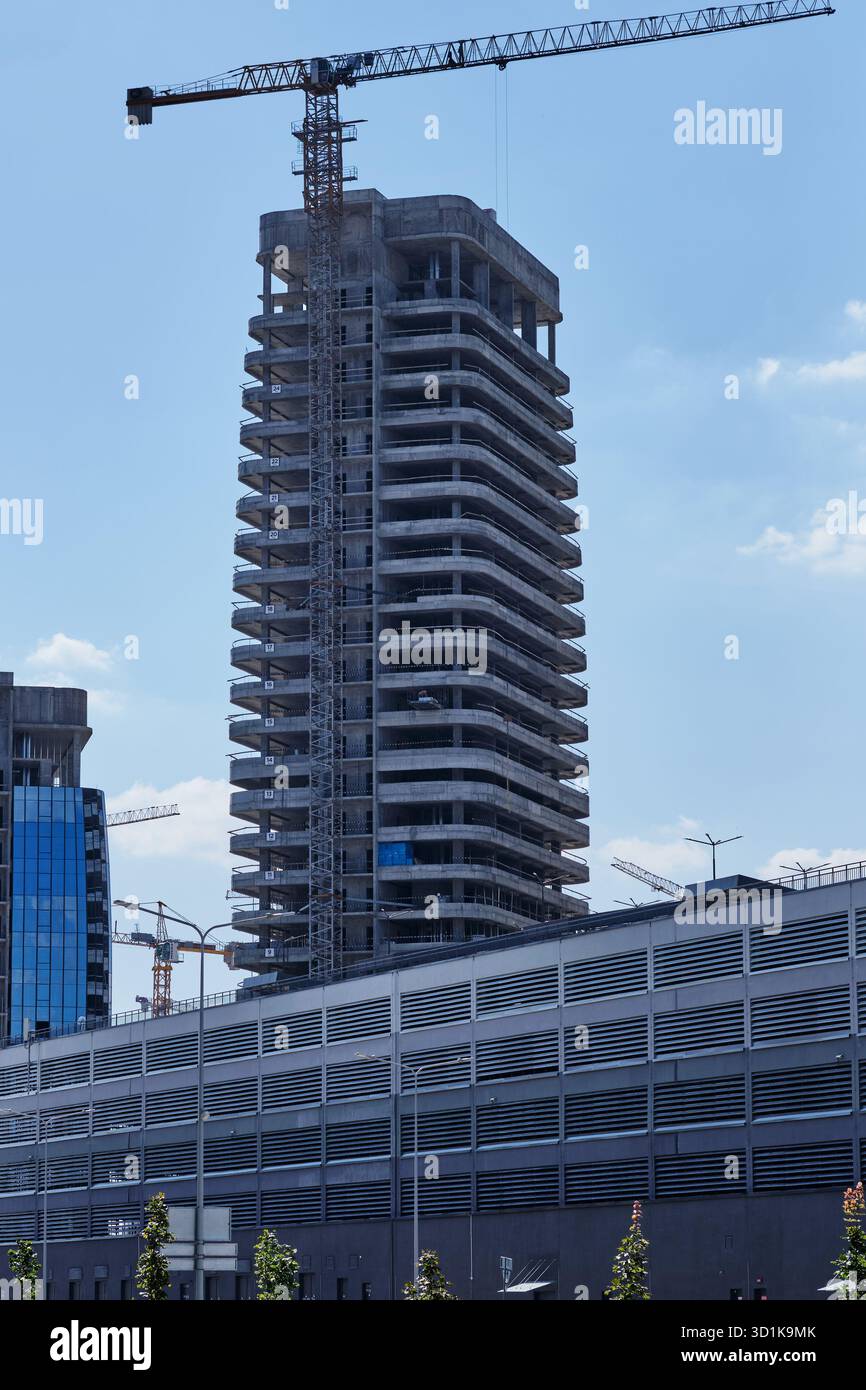 Un grattacielo in costruzione raggiunge il cielo blu, una gru arroccata sulla sua struttura scheletrica in un paesaggio urbano durante il giorno. Foto Stock