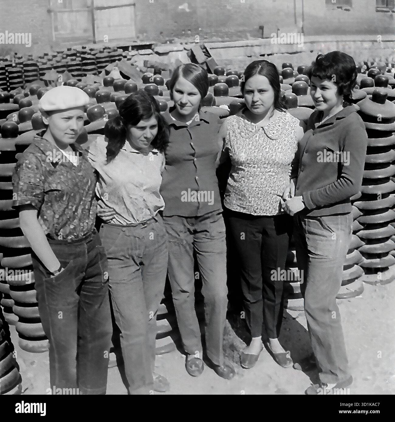 URSS, RSS Ucraina, anni 1970 Sloviansk, Oblast' di Donetsk. Impianto di isolamento e armature di Sloviansk (AIZ). Un ritratto di gruppo di cinque giovani lavoratrici. Le giovani donne stanno insieme, le braccia l'una intorno all'altra, nel magazzino all'aperto della fabbrica, circondato da enormi pile di isolanti finiti ad alta tensione. Sono vestiti in stile casual anni '1970, compresi pantaloni e berretto. Questa foto archivistica in bianco e nero cattura un momento di gioventù sovietica e cameratismo in un ambiente industriale. Foto Stock