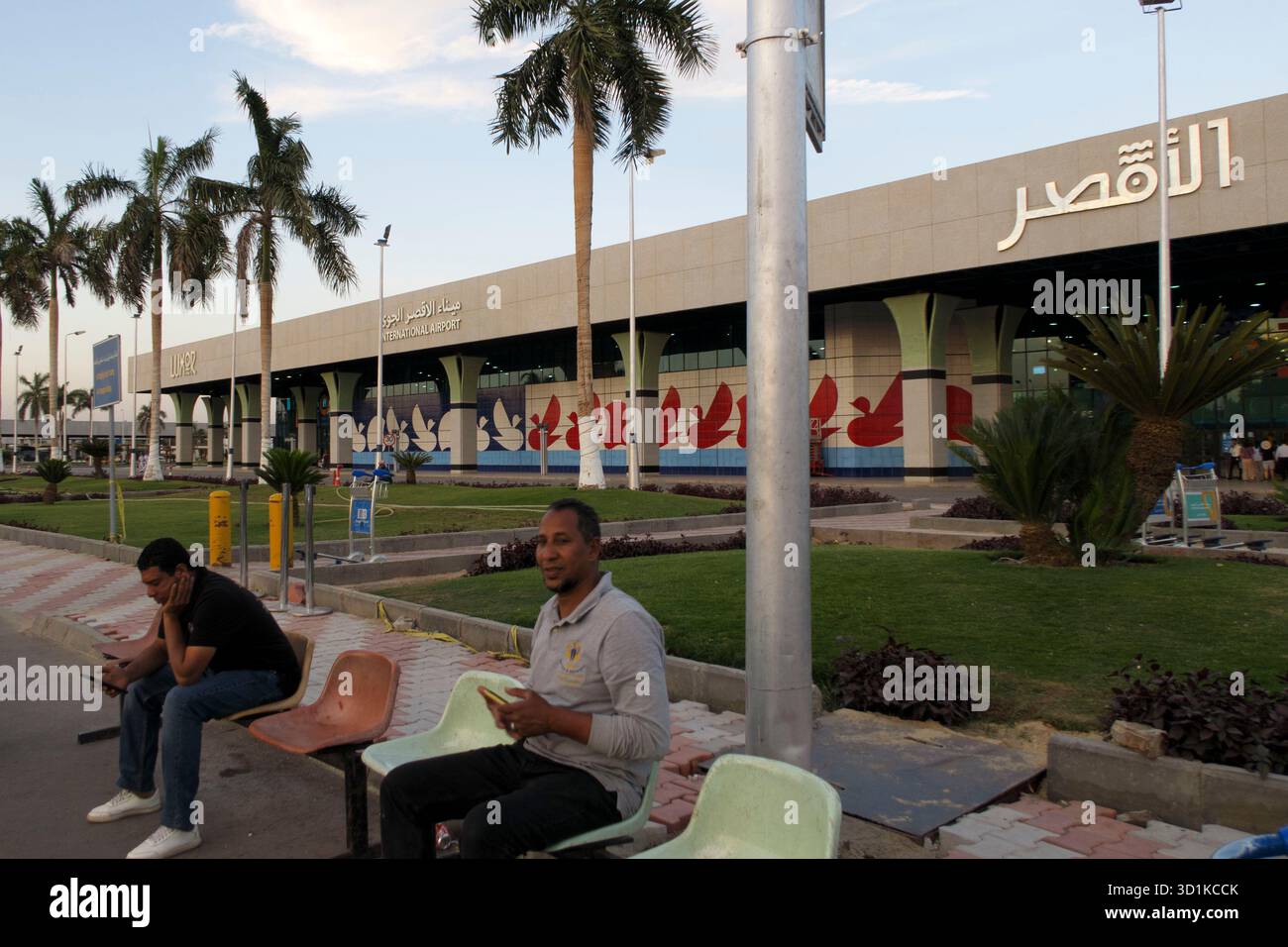 Persone sedute fuori dall'entrata dell'Aeroporto Internazionale di Luxor con palme e facciata moderna in vista, Luxor, Egitto Foto Stock