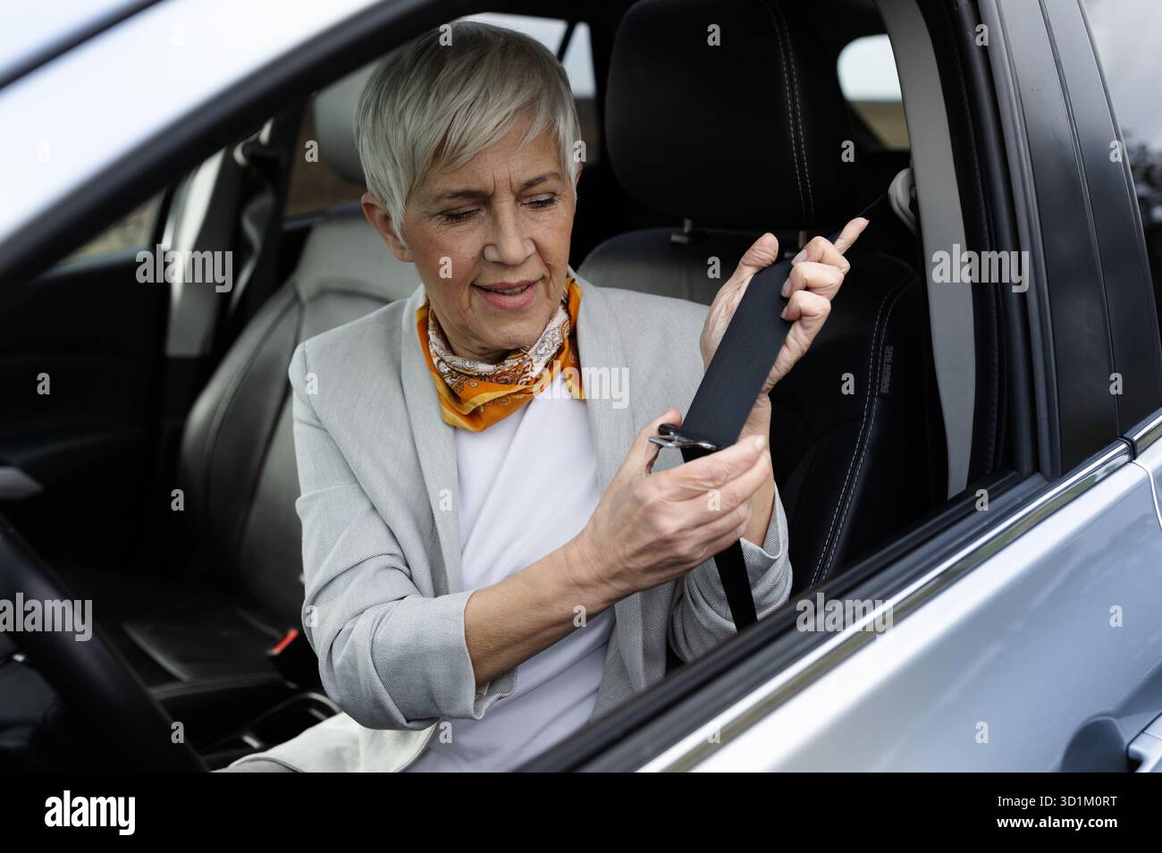 Una donna anziana siede in auto e fissa la cintura di sicurezza mentre si prepara a guidare, dimostrando consapevolezza della sicurezza e disponibilità per il viaggio che ci attende. Foto Stock