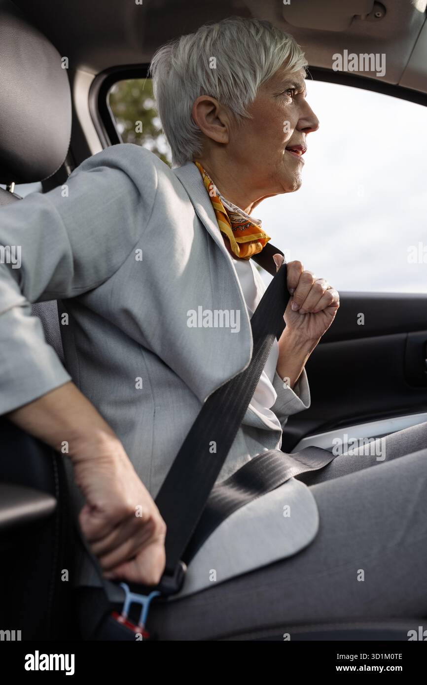 Una donna anziana siede in auto e fissa la cintura di sicurezza mentre si prepara a guidare, dimostrando consapevolezza della sicurezza e disponibilità per il viaggio che ci attende. Foto Stock