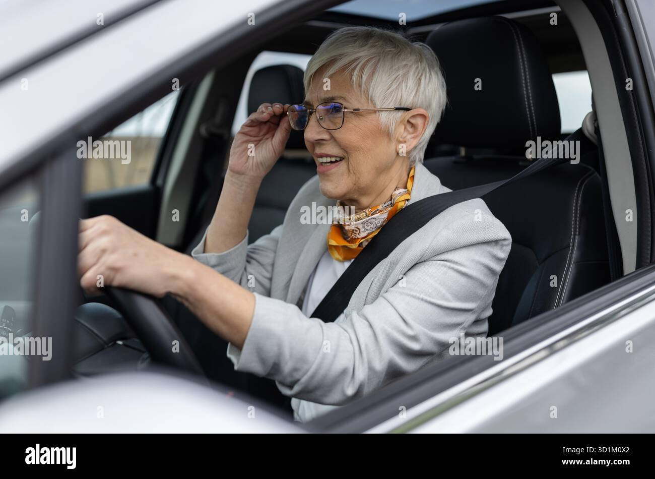 Donna anziana in abiti da lavoro che guida un'auto moderna e adatta la sua vista Foto Stock