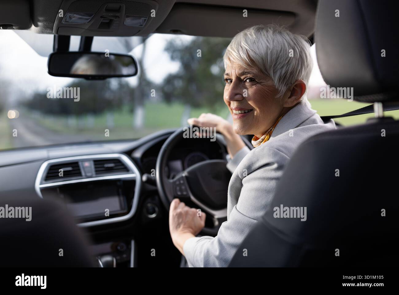 Una donna allegra ama guidare guardando indietro la fotocamera. Si trova all'interno di un'auto moderna con sfondo panoramico Foto Stock