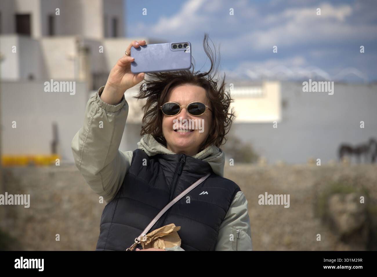 Una donna sorridente che indossa gli occhiali da sole e una giacca a piumino bicolore regge uno smartphone per scattare un selfie. I suoi capelli sono travolti dal vento Foto Stock