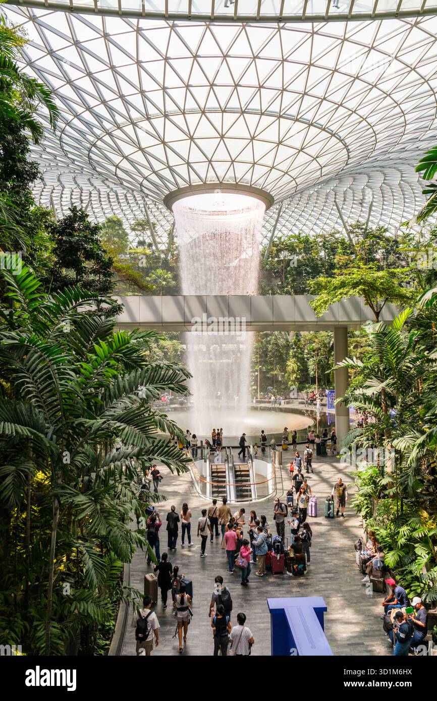 Jewel Changi Airport mette in mostra lo spettacolare Rain Vortex circondato da piante tropicali, con i viaggiatori che esplorano lo spazio vivace. Foto Stock