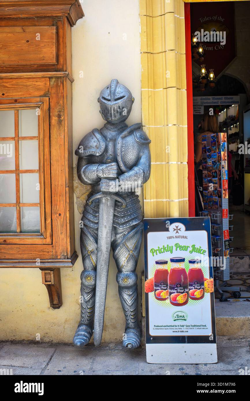 Mdina, Malta - 09.20.2025: Una statua di un cavaliere medievale in armatura si trova all'ingresso di un negozio di souvenir accanto a un poster di succo di fico d'India. Foto Stock