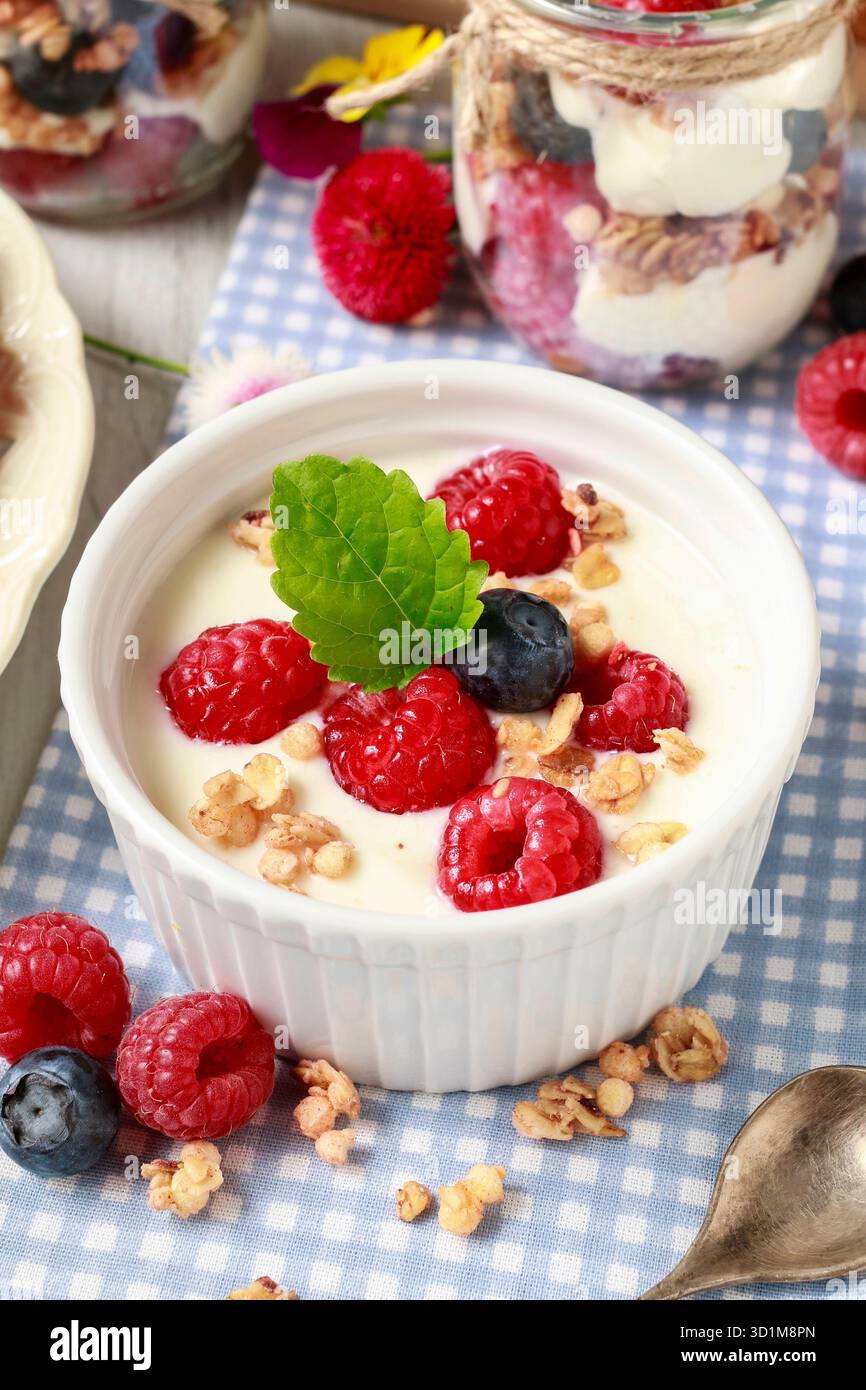 Tavolo della colazione: Ciotola di yogurt con muesli e frutta fresca: Lamponi e mirtilli, vasetti di vetro con frutta e panna montata. Foto Stock
