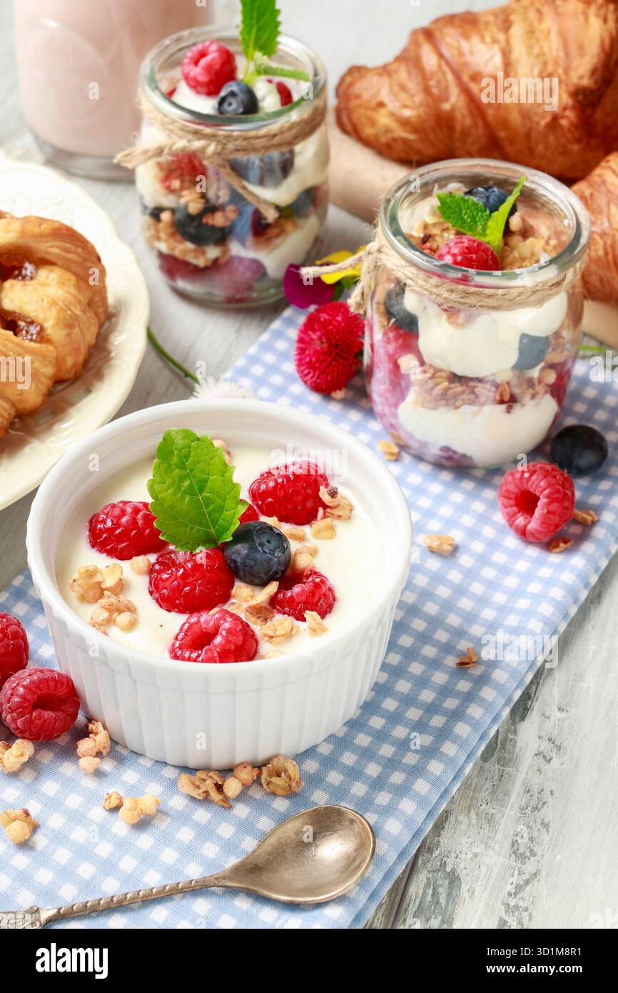 Tavolo della colazione: Ciotola di yogurt con muesli e frutta fresca: Lamponi e mirtilli, vasetti di vetro con frutta e panna montata. Foto Stock