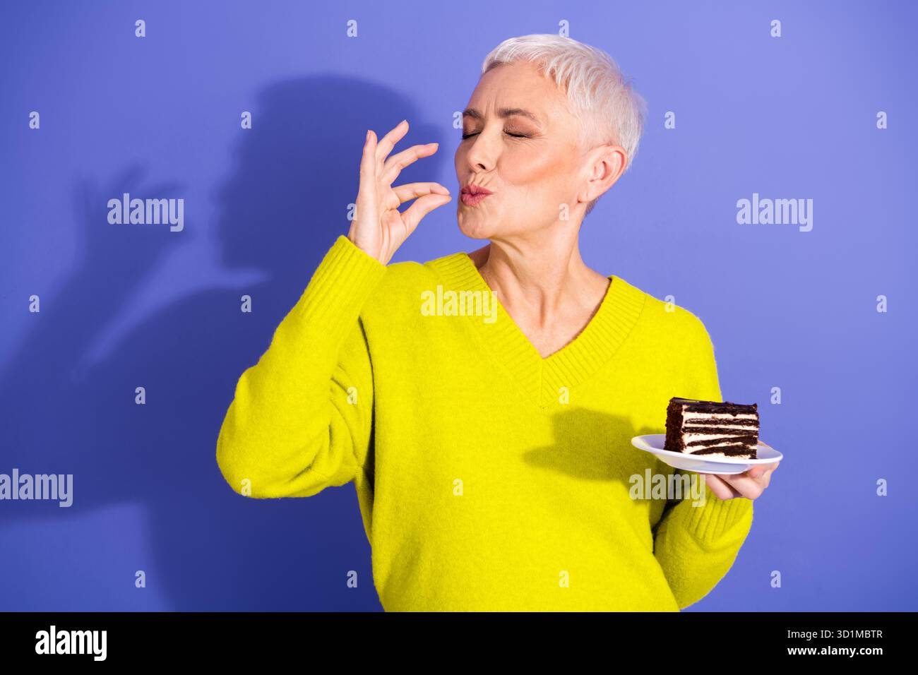 Una donna anziana con maglione giallo brillante si gode una torta e un bacio giocoso su sfondo viola in un momento di vita gioioso Foto Stock