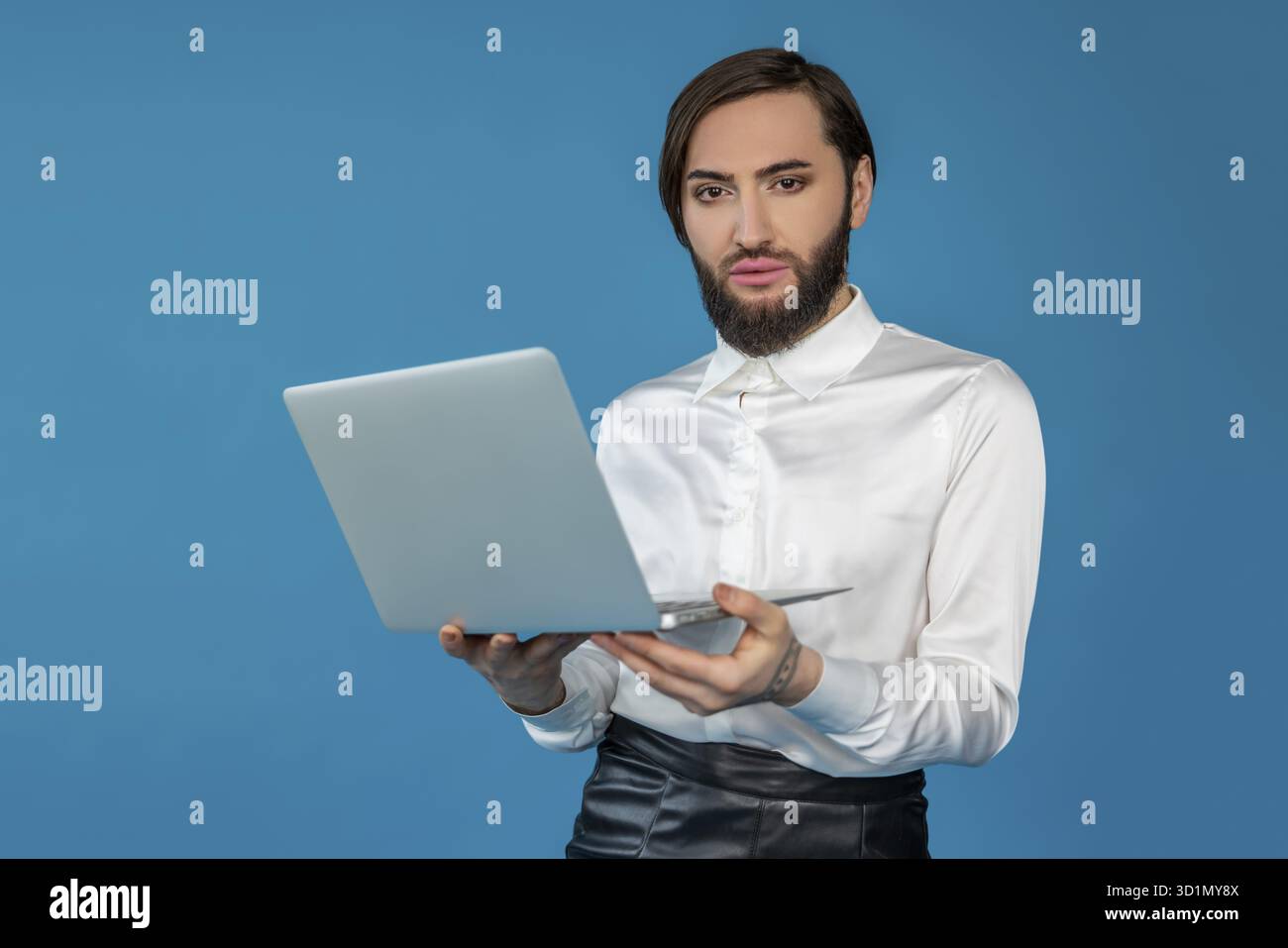 Uomo transgender sicuro di sé che indossa una camicia bianca che lavora su un laptop che naviga su Internet e guarda la fotocamera che posa isolata su sfondo blu Foto Stock