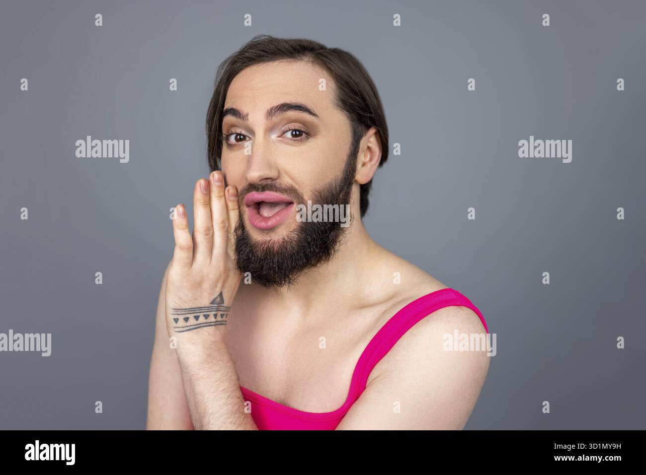 Una bruna allegra e ottimista transessuale con la parte superiore rosa tiene la mano vicino alla bocca dicendo qualcosa che condivide la posa segreta isolata su sfondo grigio Foto Stock
